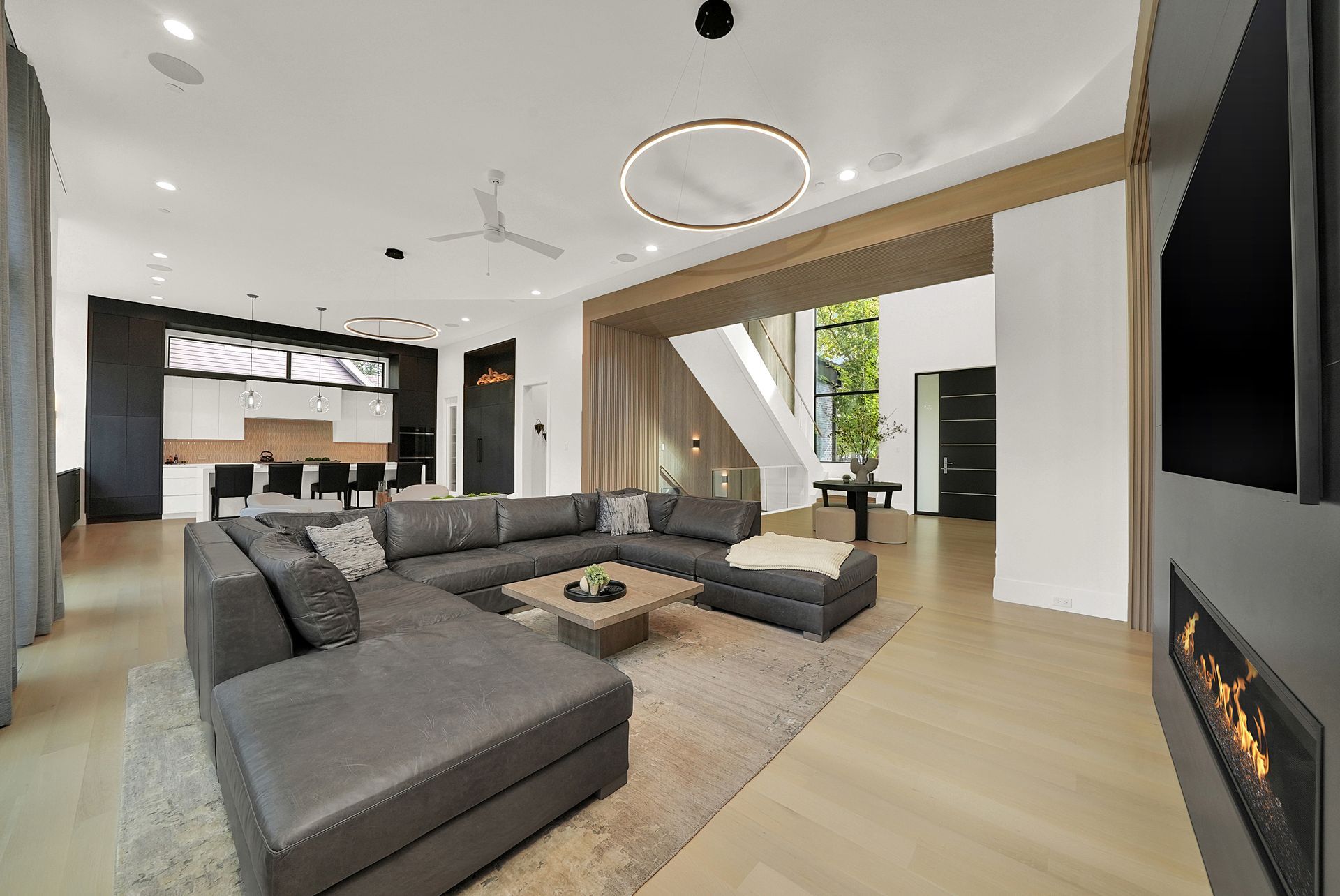 Modern open-plan living room with gray sectional sofa, fireplace, large TV, and floor-to-ceiling windows