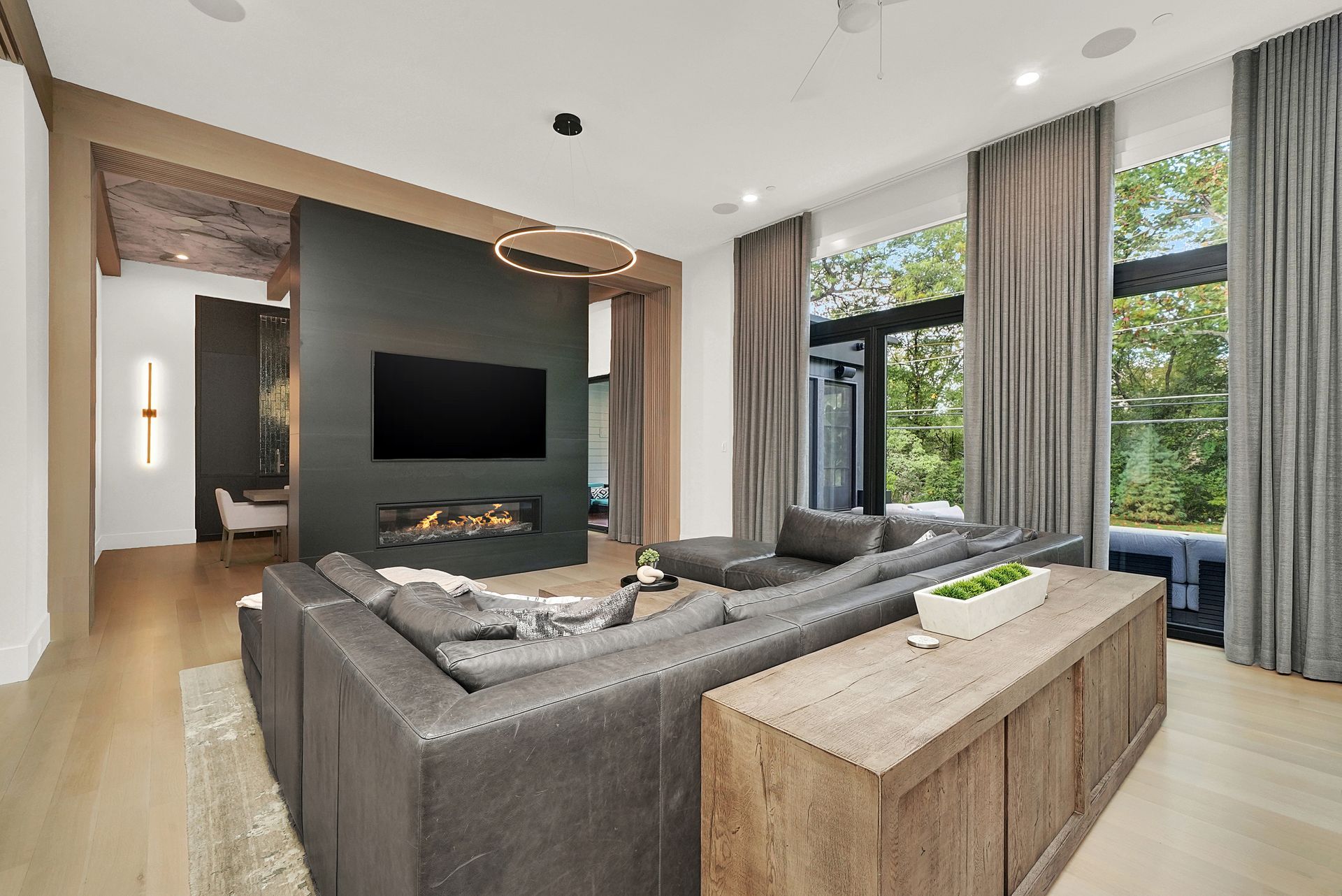 Modern living room with gray sectional, wall-mounted TV, fireplace, and large windows overlooking greenery