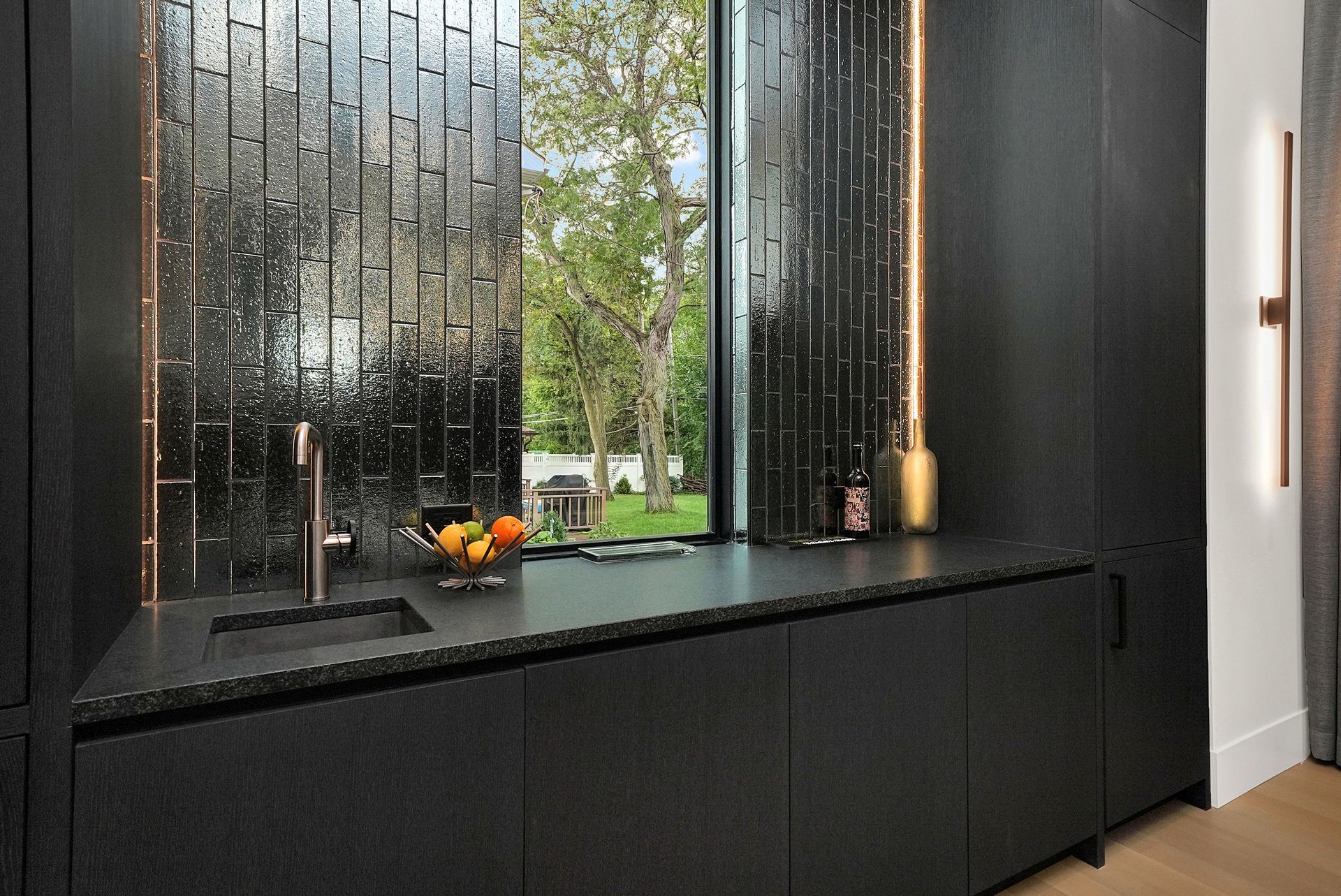 Modern black kitchen counter with sink, fruit bowl, and textured backsplash facing a window view of trees.