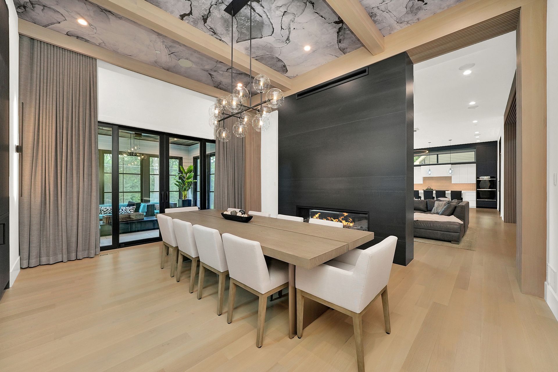 Modern dining room with long wooden table, white chairs, tall curtains, and black accent wall in open interior