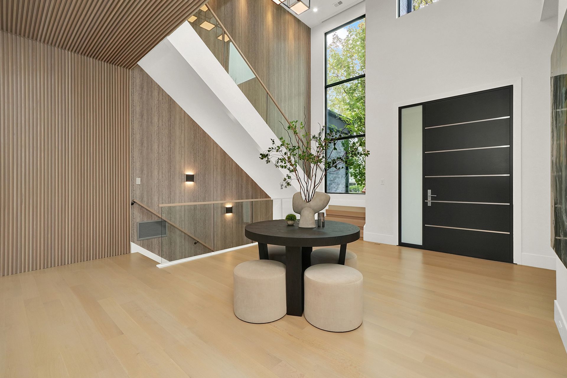 Modern foyer with light wood floors, black front door, round table, two stools, and a staircase with a glass railing