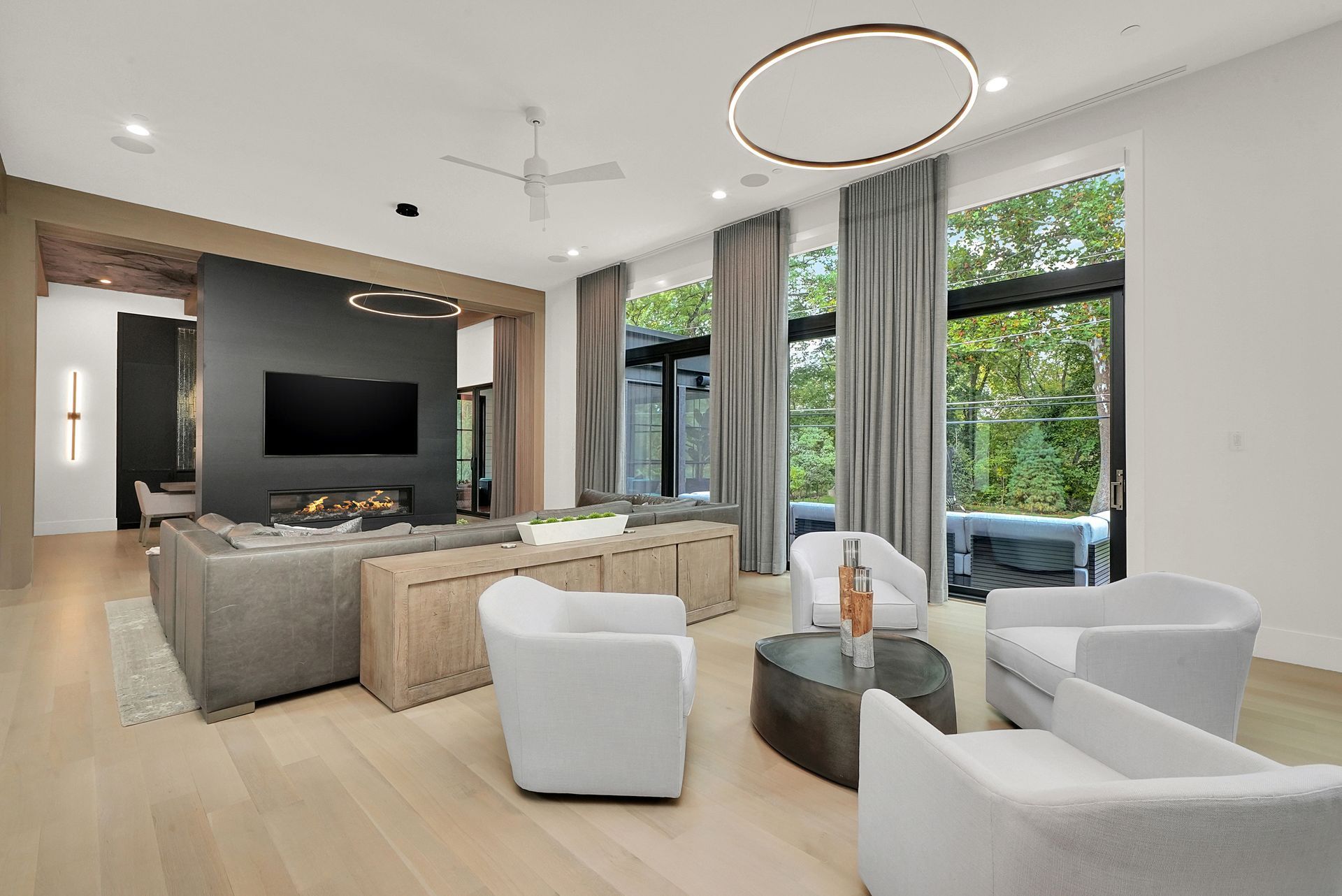 Modern living room with gray sofas, white chairs, large TV, and floor-to-ceiling windows