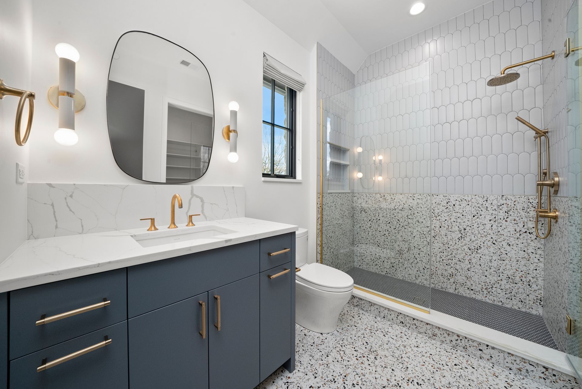Modern bathroom with blue vanity, gold fixtures, white tile shower, and speckled terrazzo floor