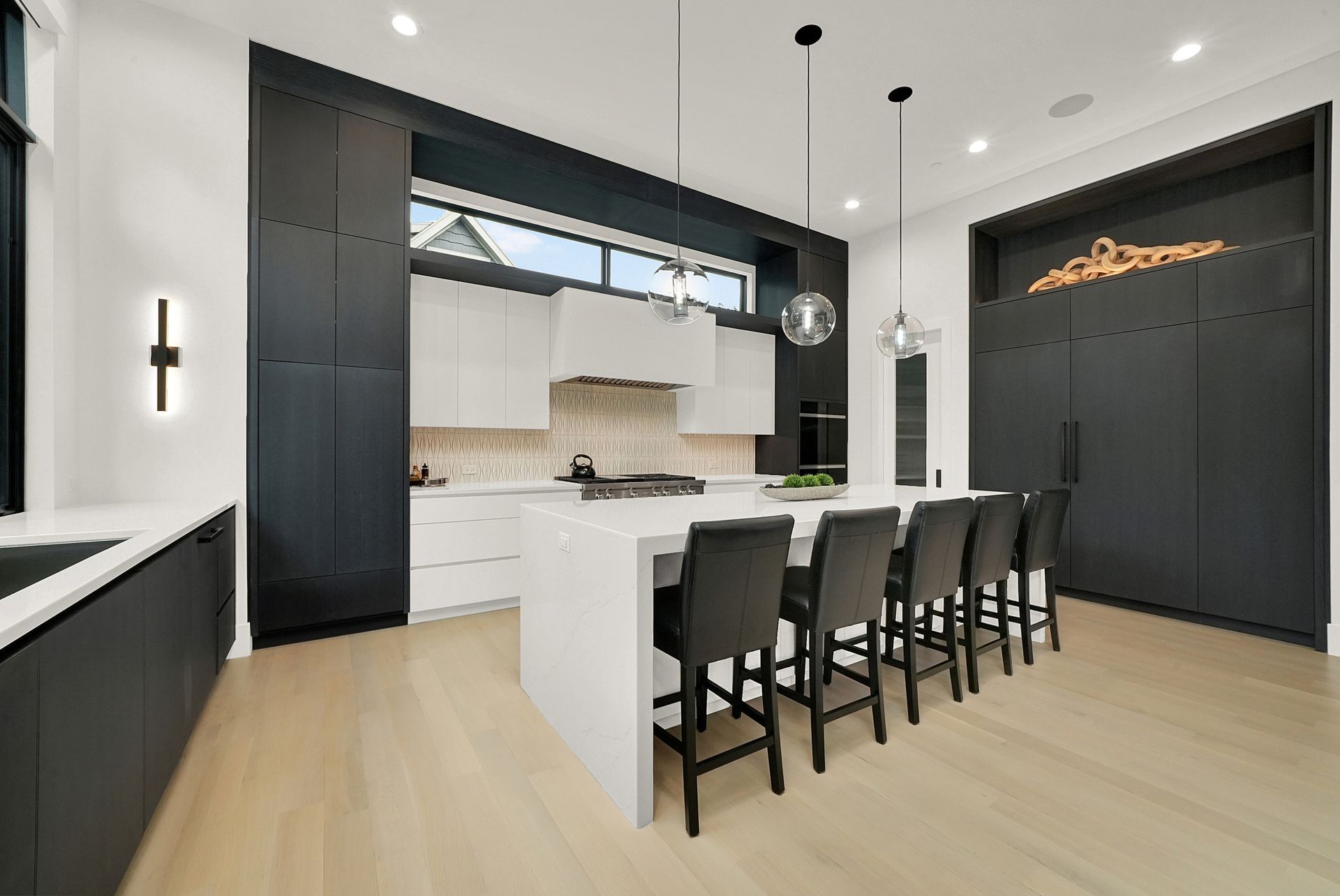 Modern kitchen with black island chairs, light wood floors, and dark cabinetry under pendant lights