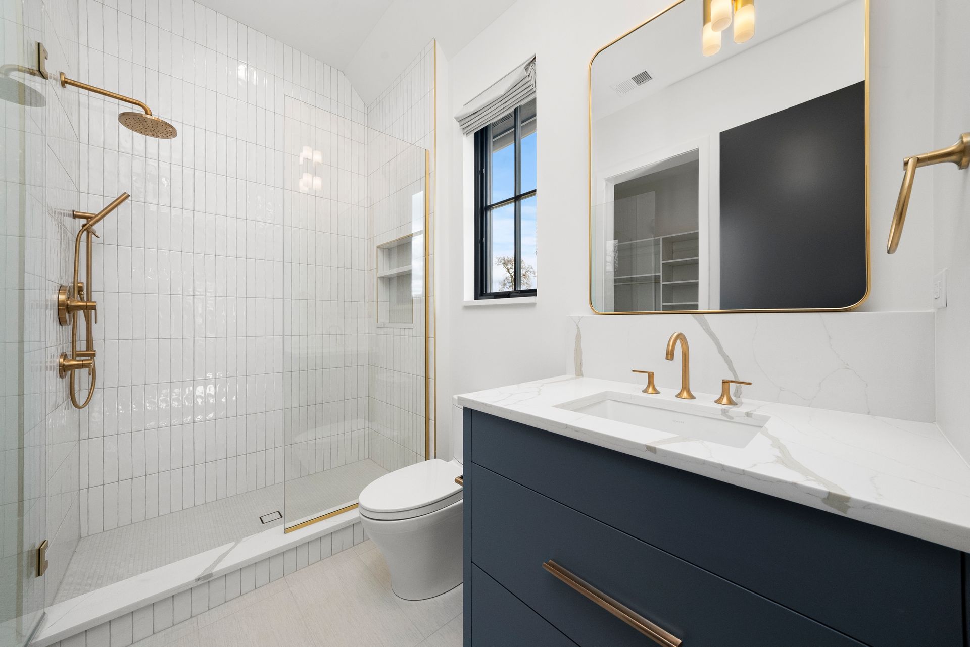 Modern white bathroom with glass shower, navy vanity, gold fixtures, and a window.