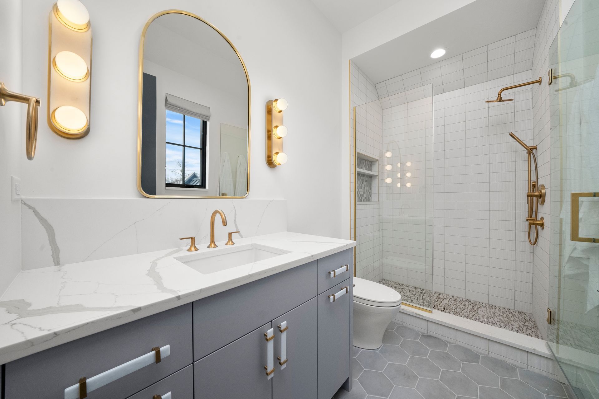 Modern bathroom with a gray vanity, gold fixtures, arched mirror, and glass shower.