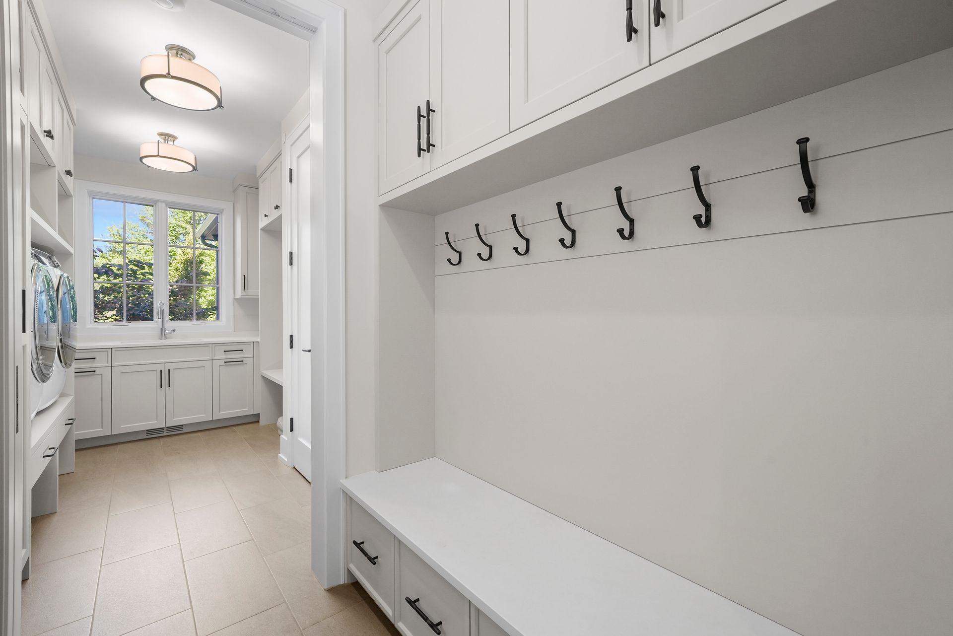 White mudroom with built-in cabinets, bench, hooks, and a bright window at the end of the hall.