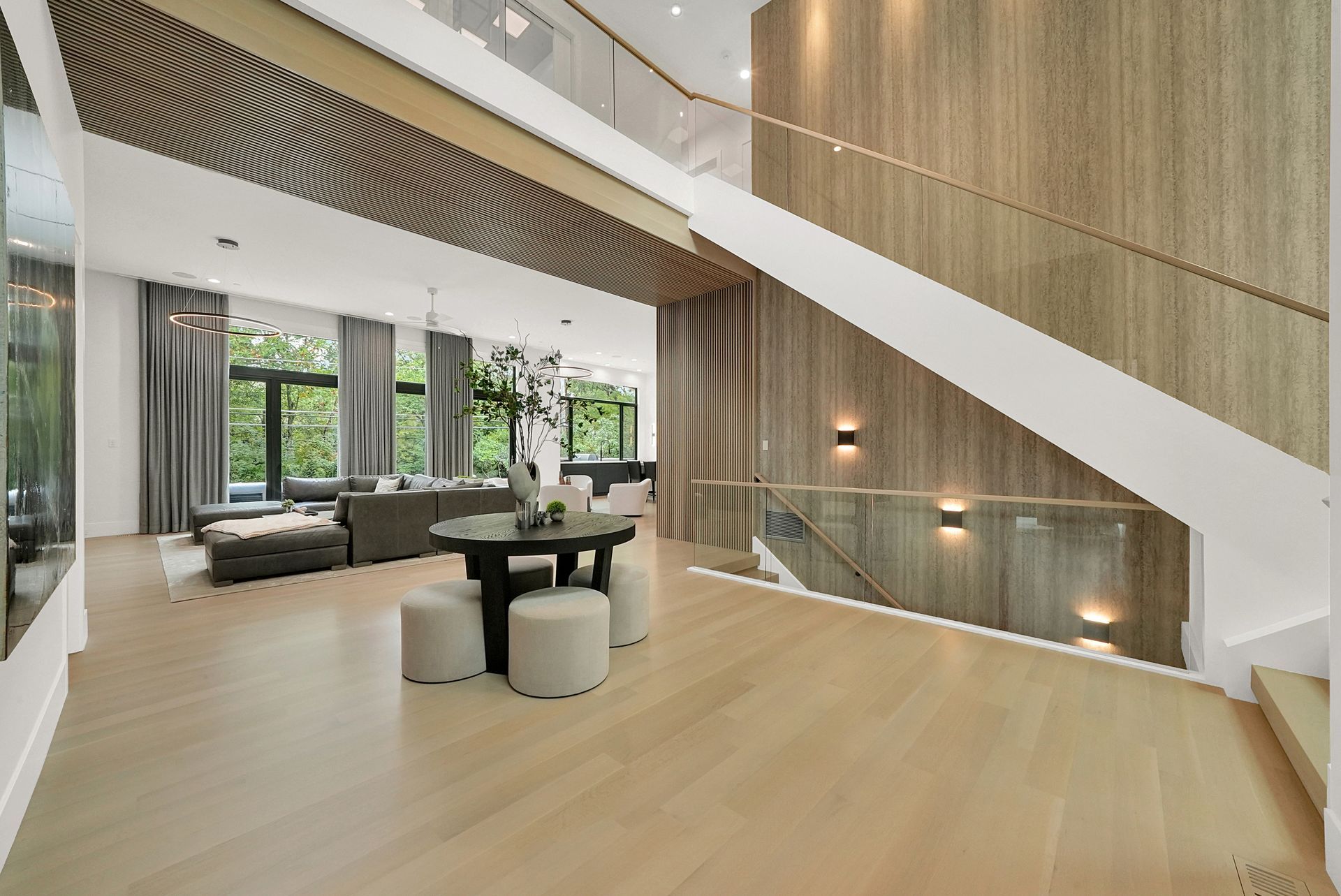 Modern open-concept living room with light wood floors, large windows, and a staircase with glass railing