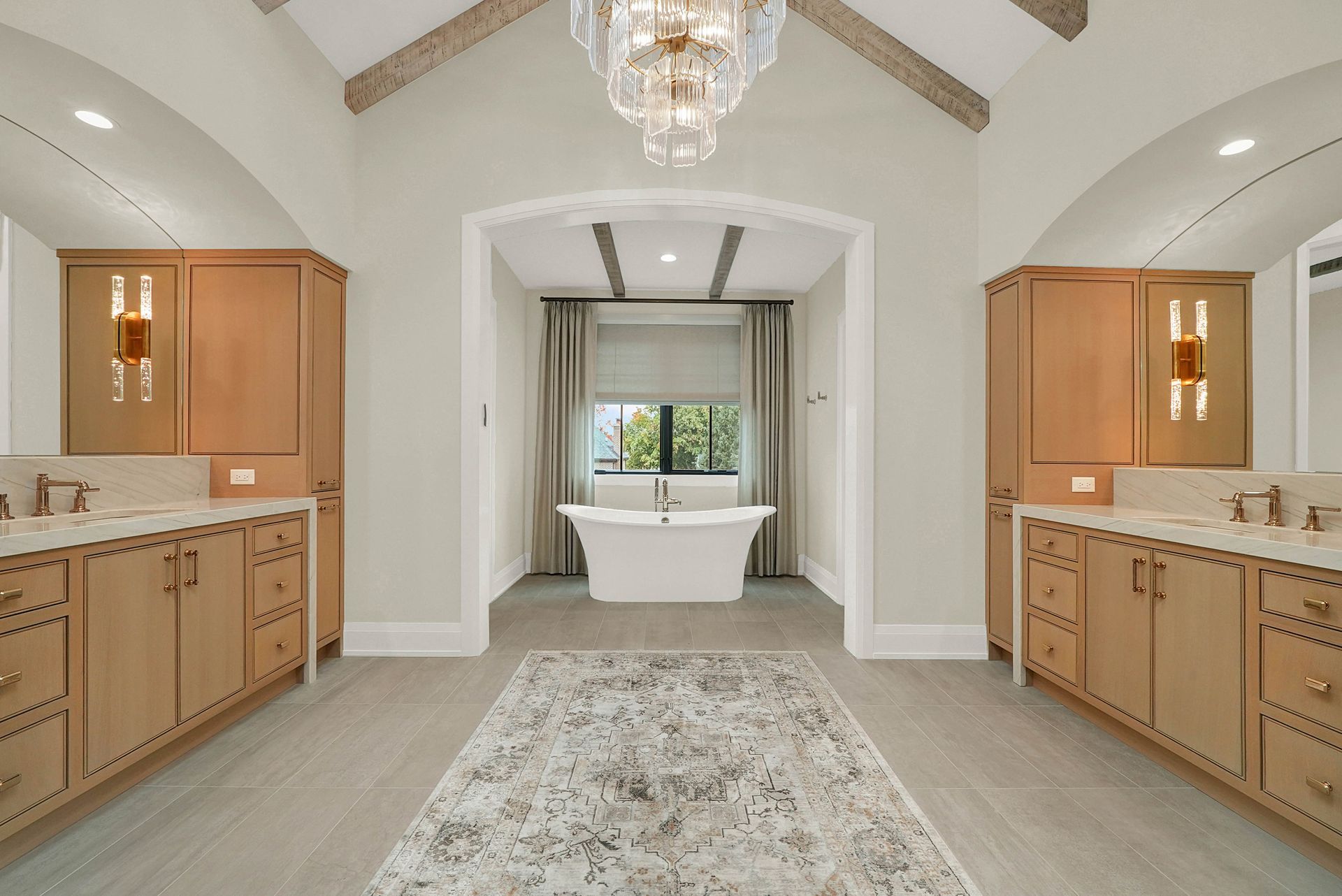 Elegant bathroom with double vanities, chandelier, and a freestanding tub under a window.