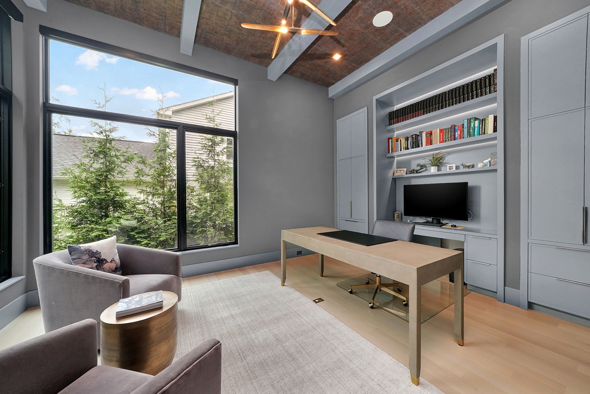 Modern living room with large windows, gray sofa, wooden coffee table, and built-in shelving with TV