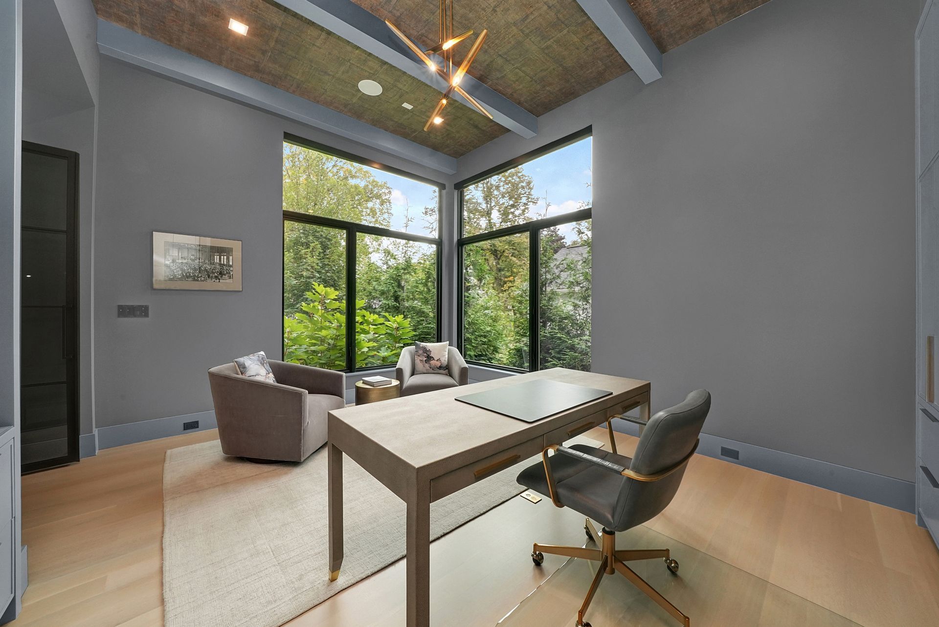 Modern home office with desk, gray chair, sofa, and large windows overlooking greenery