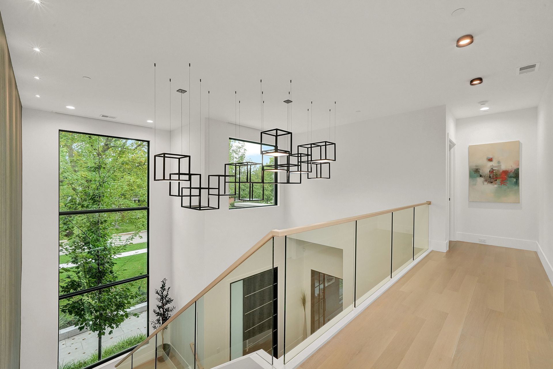 Modern white staircase landing with glass railing, light wood floors, and geometric pendant lights; green trees outside.
