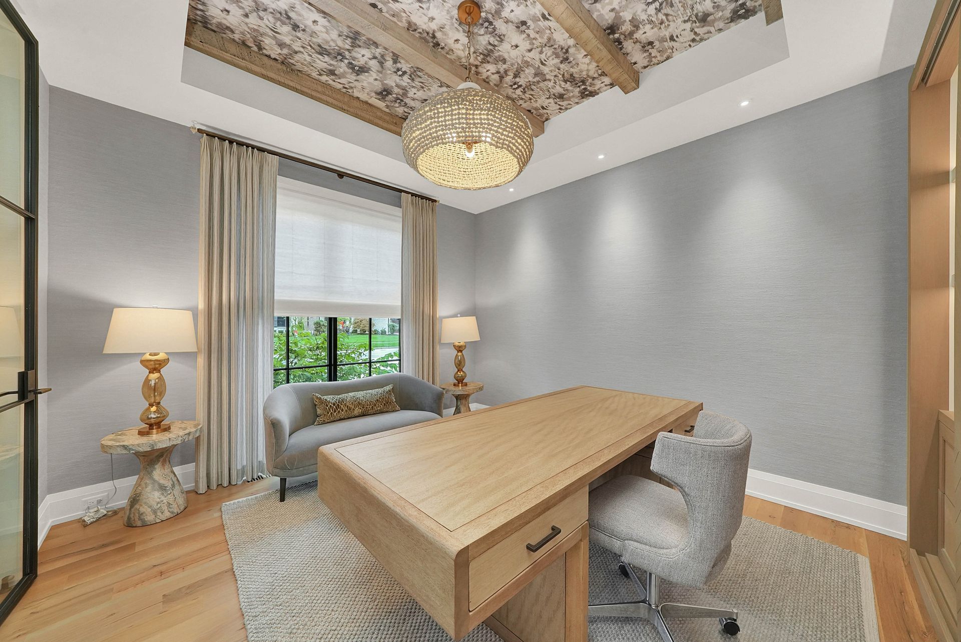 Stylish home office with wooden desk, gray chair, lamps, large window, and decorative ceiling fixture