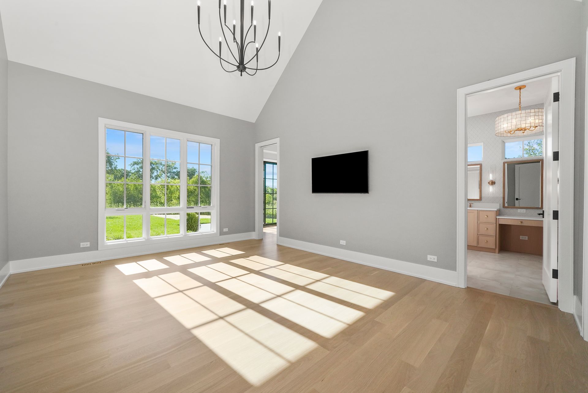 Bright empty living room with large windows, wall-mounted TV, and sunlit hardwood floors leading to a kitchen.