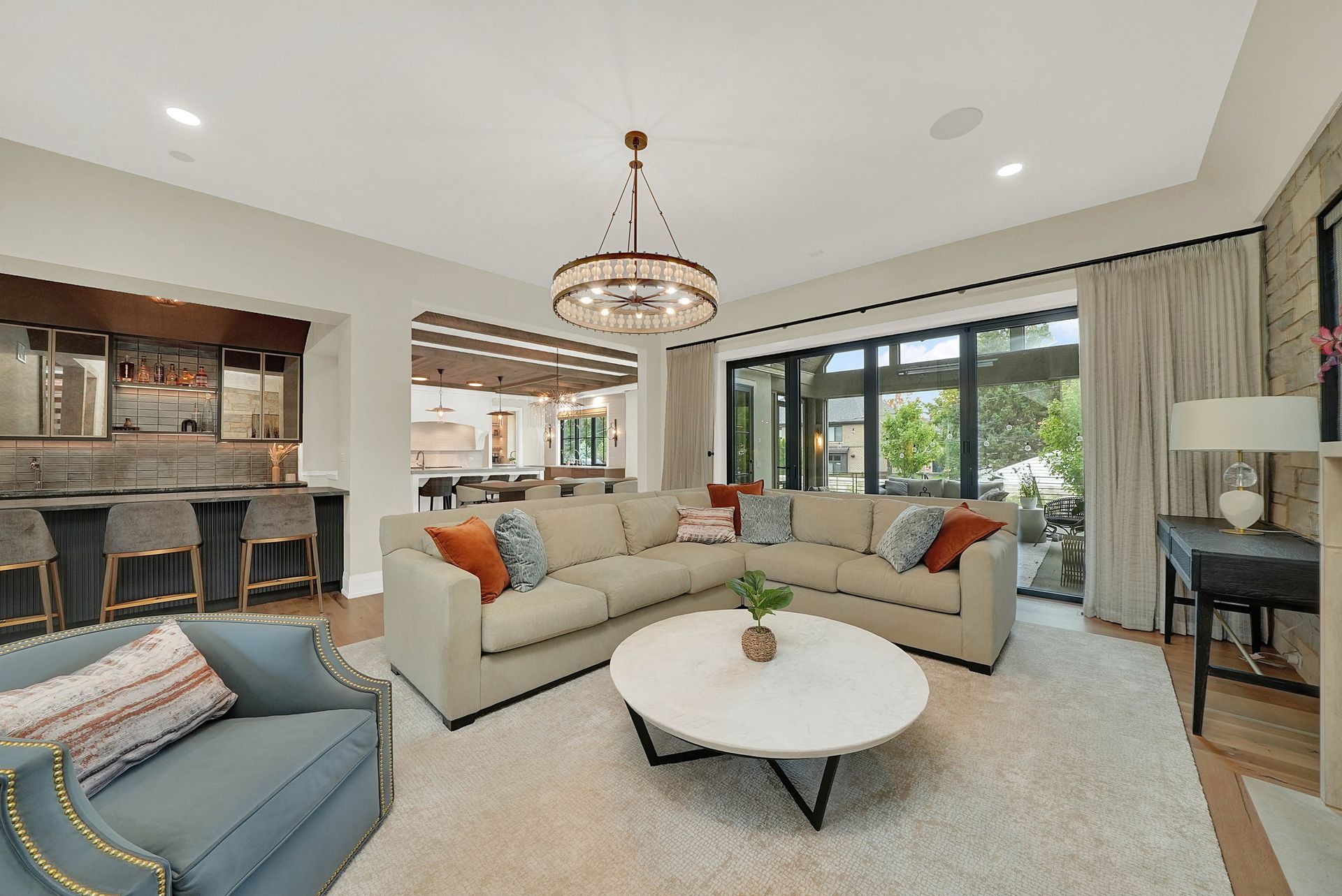 Bright living room with beige sectional, blue chair, round white coffee table, and large windows overlooking greenery