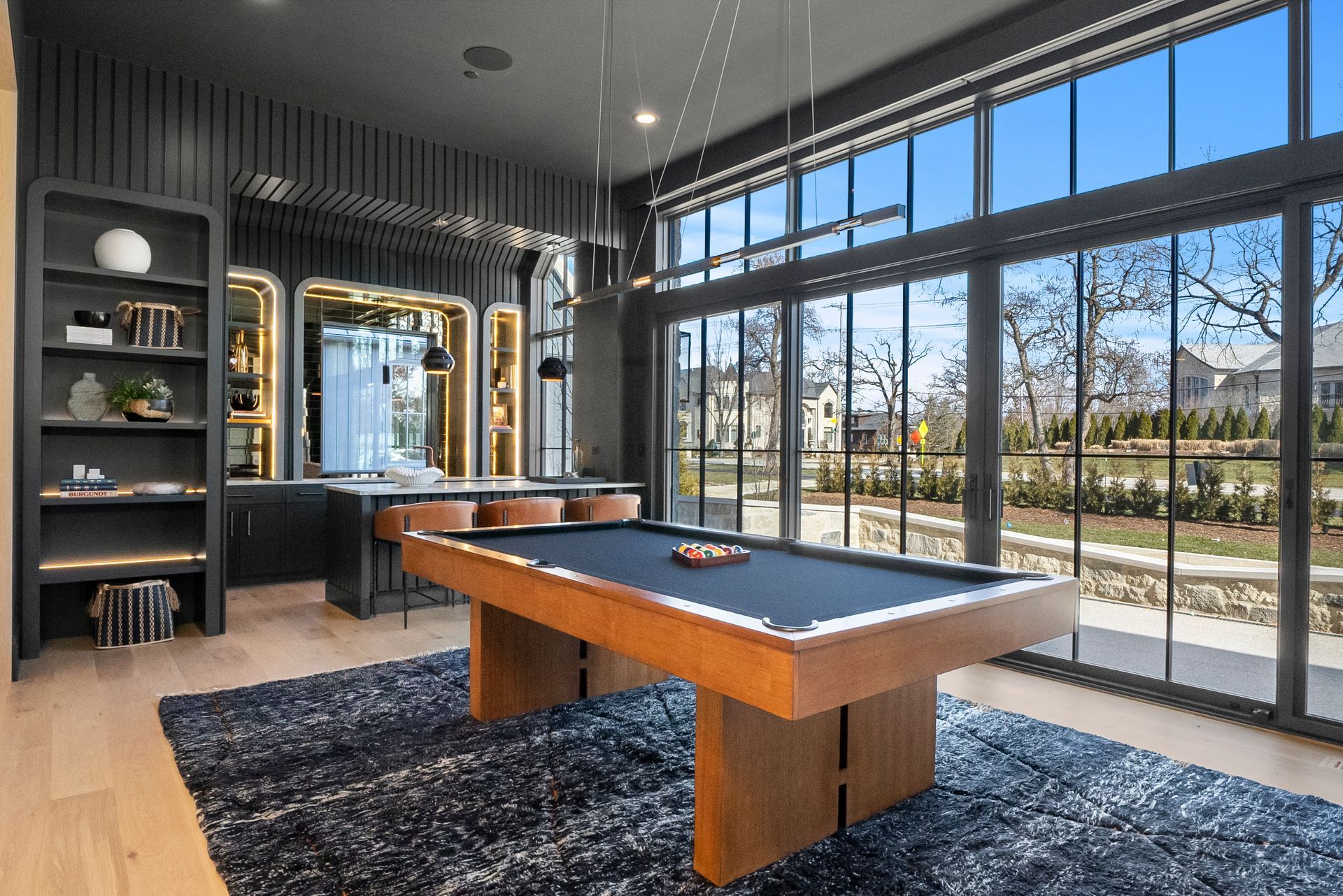 Modern game room with a pool table, dark decor, and floor-to-ceiling windows overlooking trees and a road