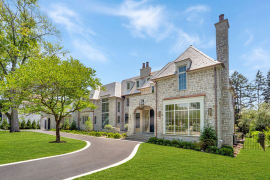 Large stone house with a manicured lawn, curved driveway, and trees under a bright sky