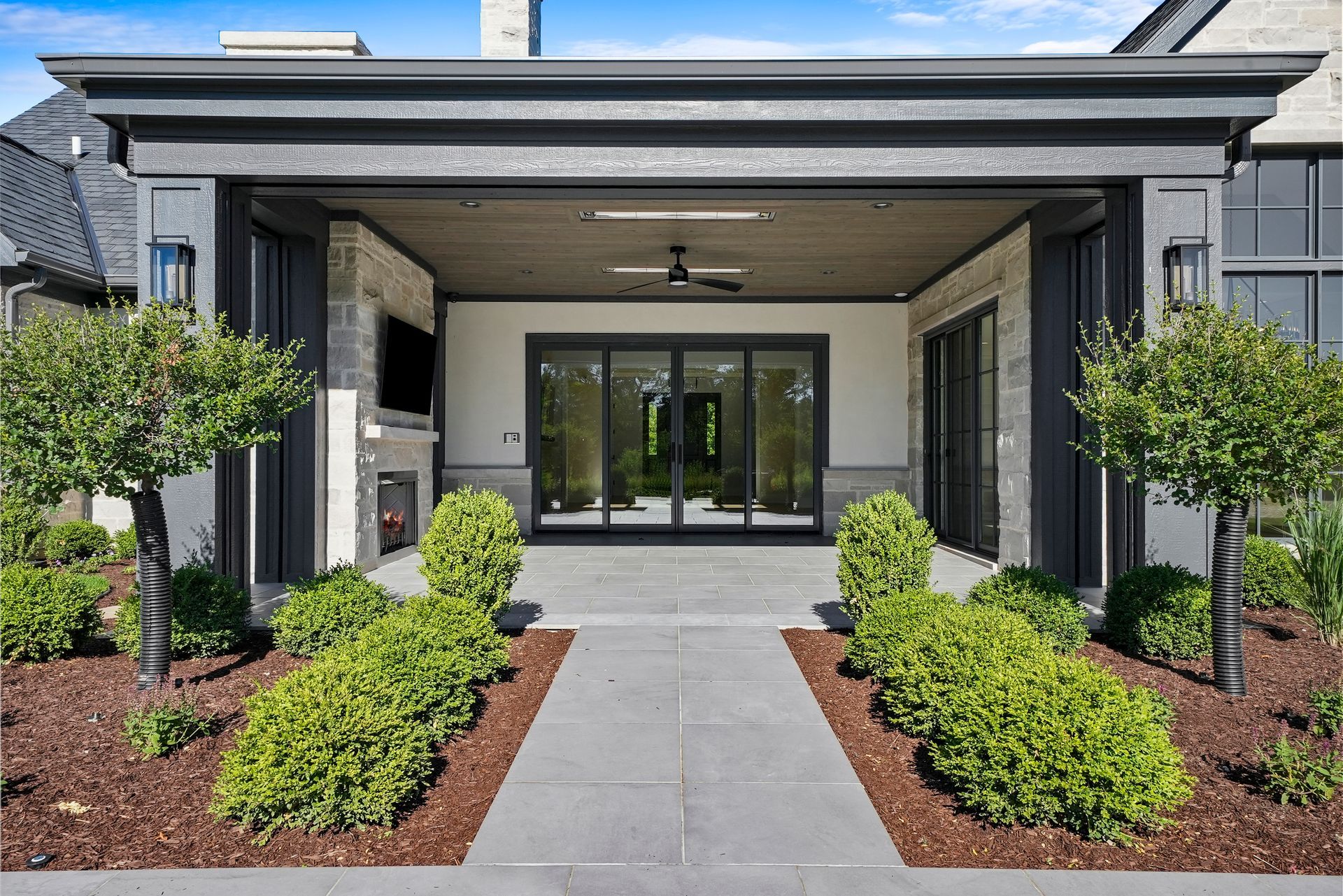 Modern home entrance with covered porch, glass doors, and landscaped shrubs along a stone walkway