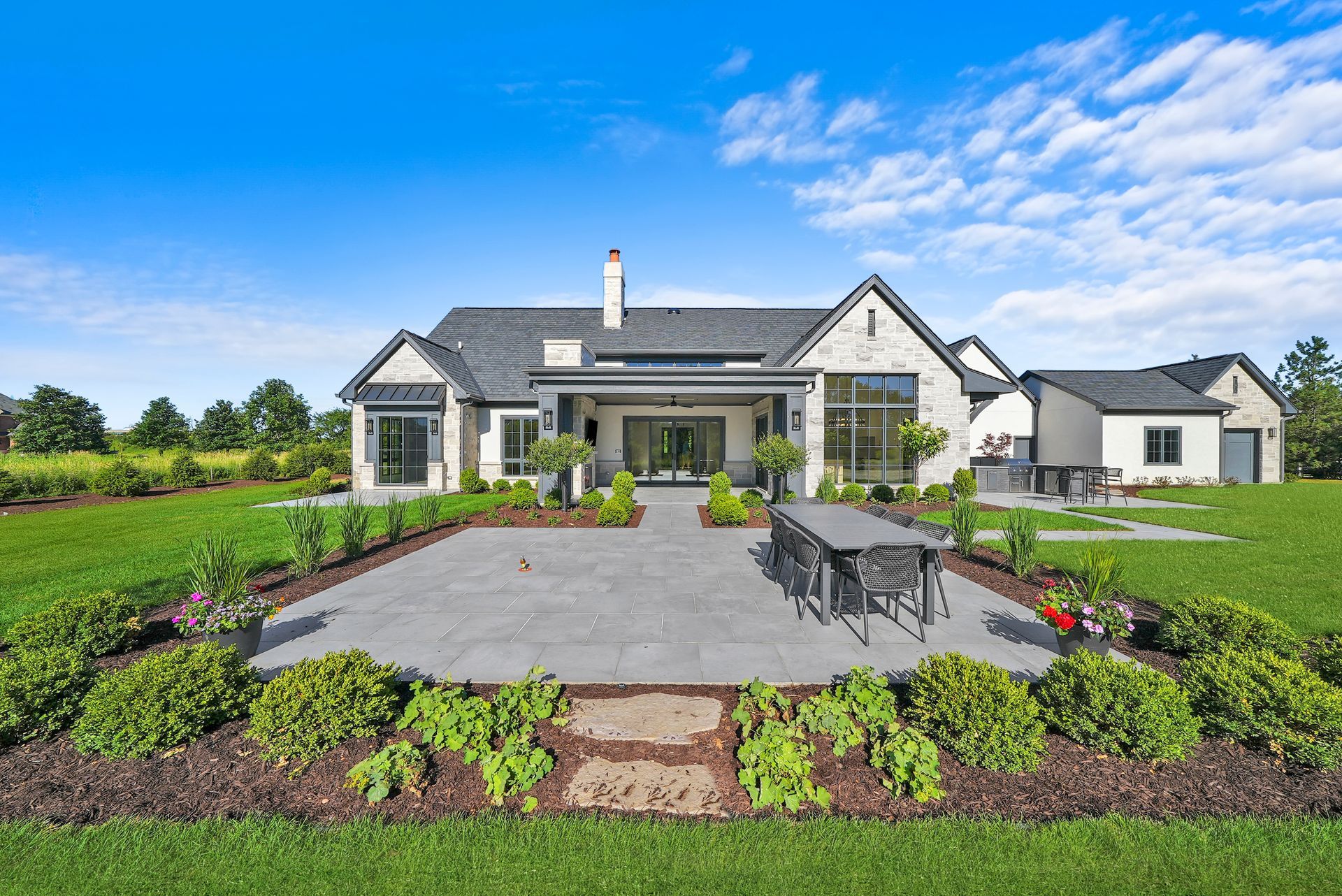 Modern house with a large paved patio, landscaped garden, and green lawn under a blue sky