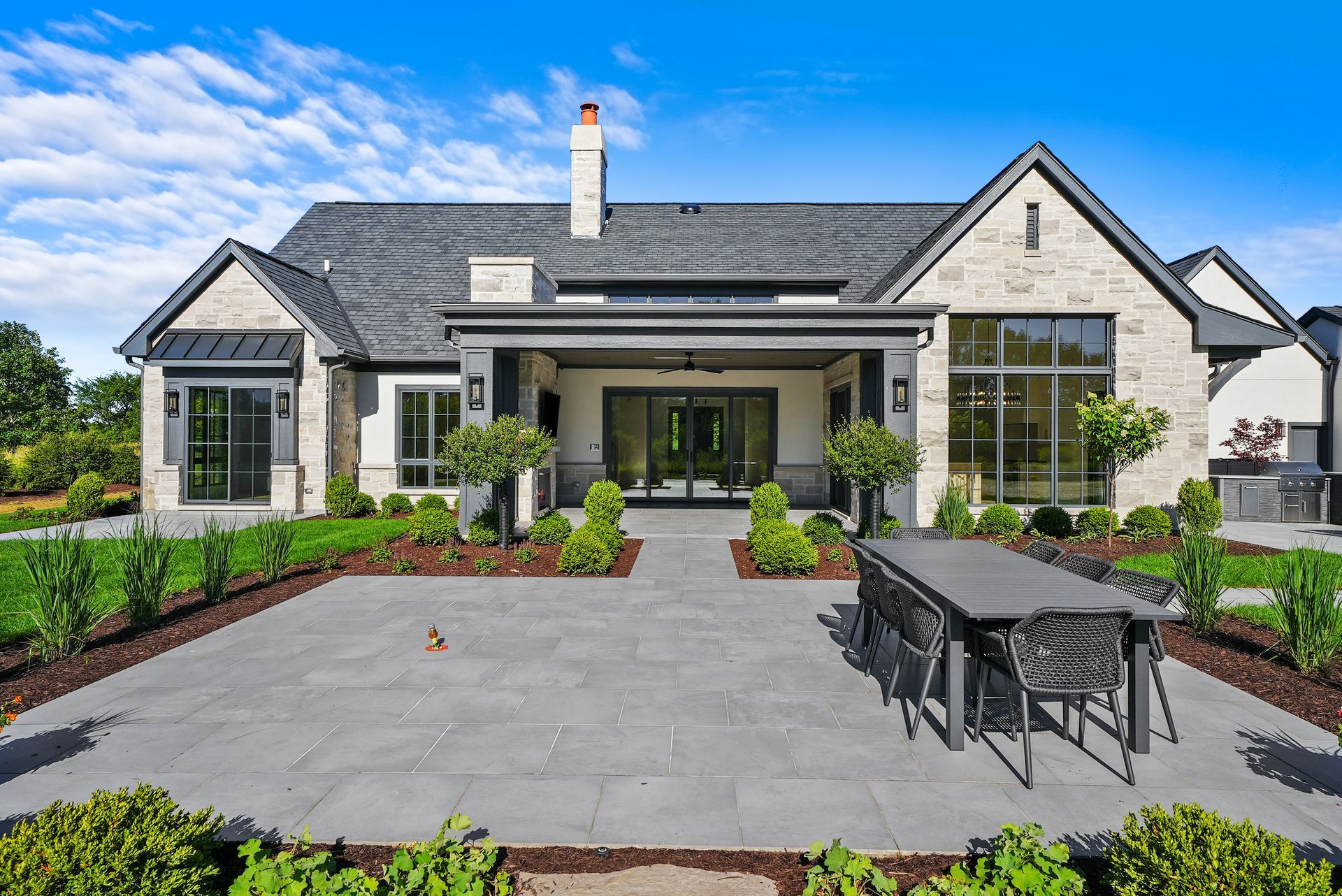 Modern single-story house with stone patio, outdoor dining set, and landscaped front yard under a blue sky
