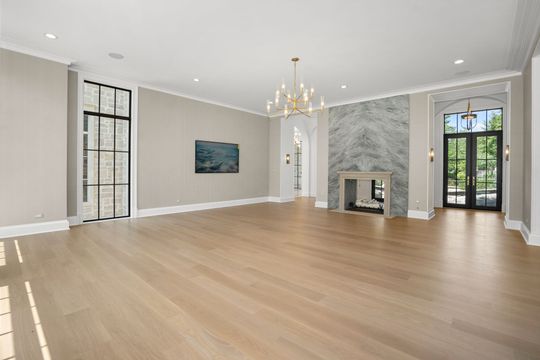 Empty modern living room with light wood floors, white walls, chandelier, fireplace, and tall windows