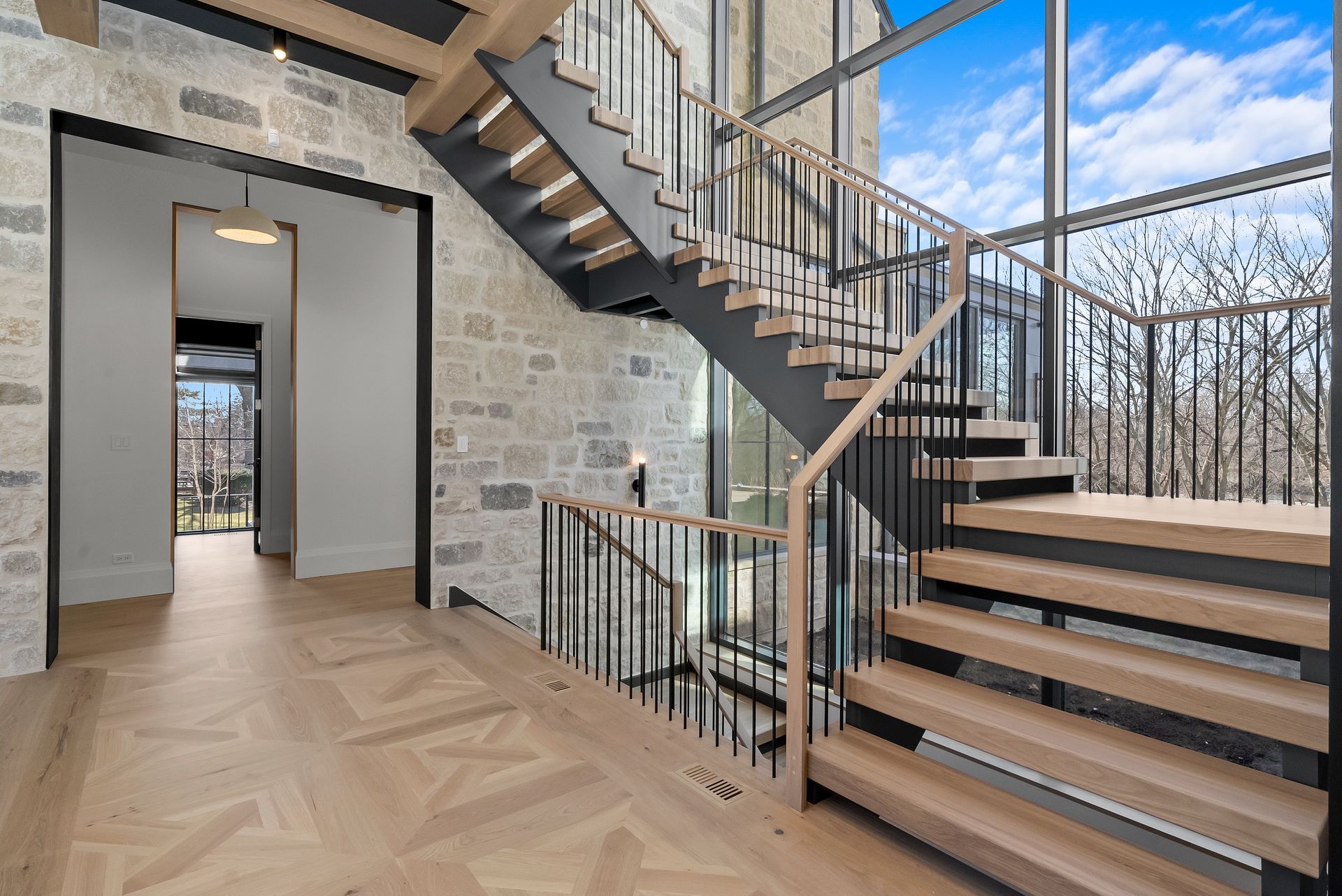 Modern staircase in a stone entryway with wood steps, metal rails, and large windows overlooking trees.