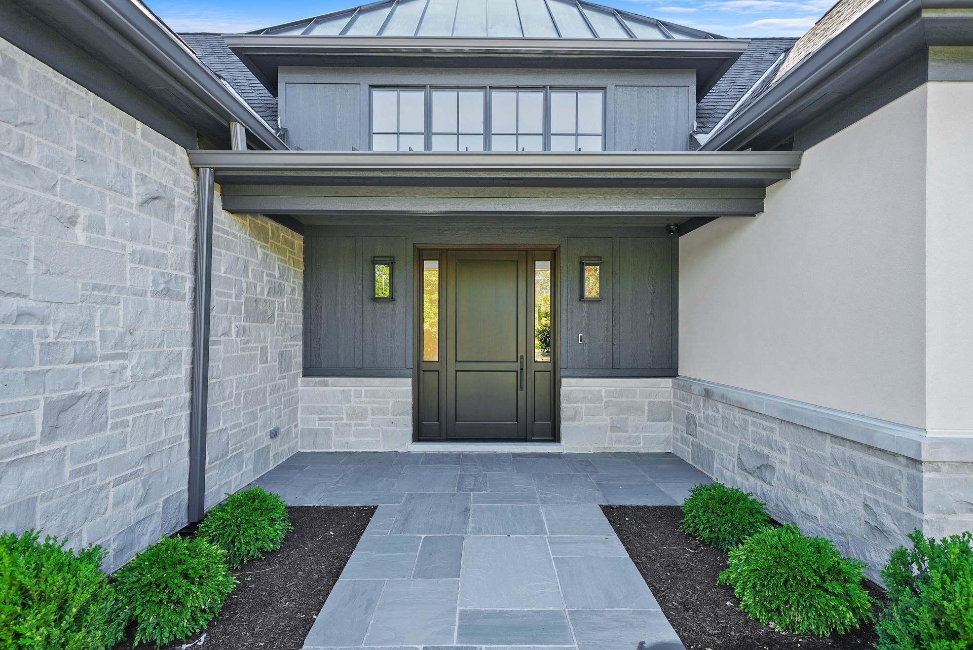 Modern home front entrance with stone walls, double doors, and a paved walkway lined with shrubs