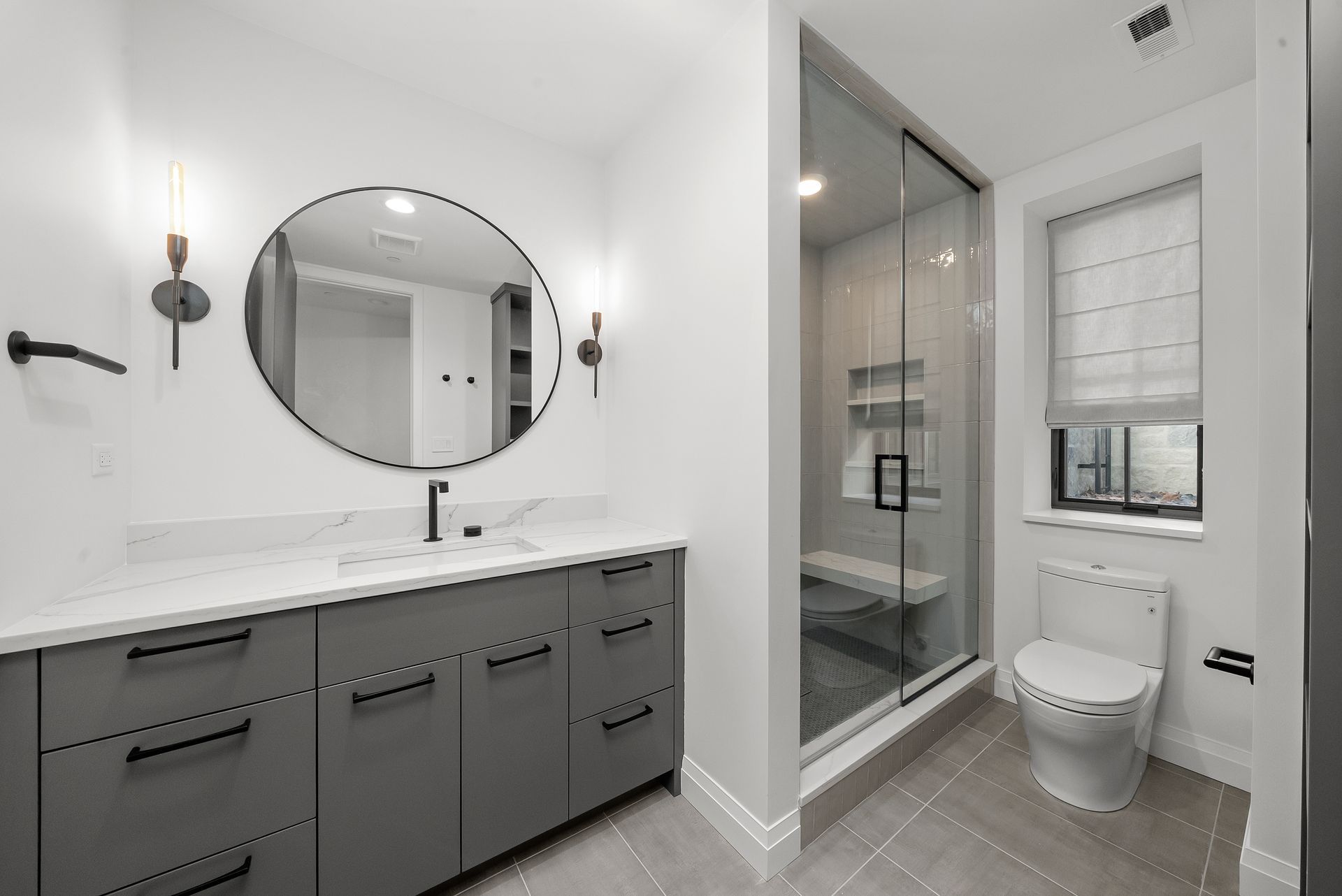 Modern bathroom with gray vanity, round mirror, walk-in shower, and toilet near a window