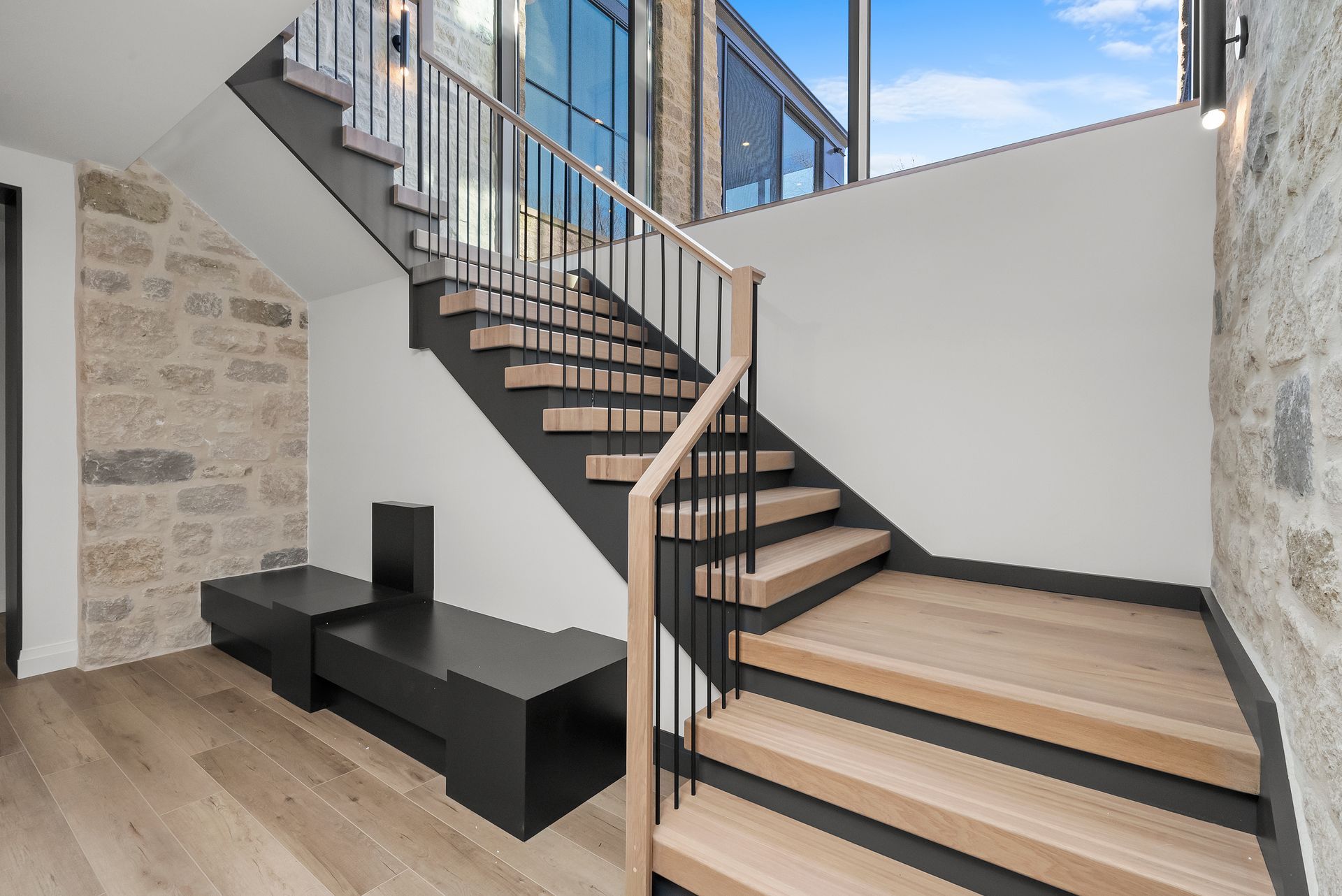 Modern staircase in a bright stone-and-glass interior with black railings and wood steps