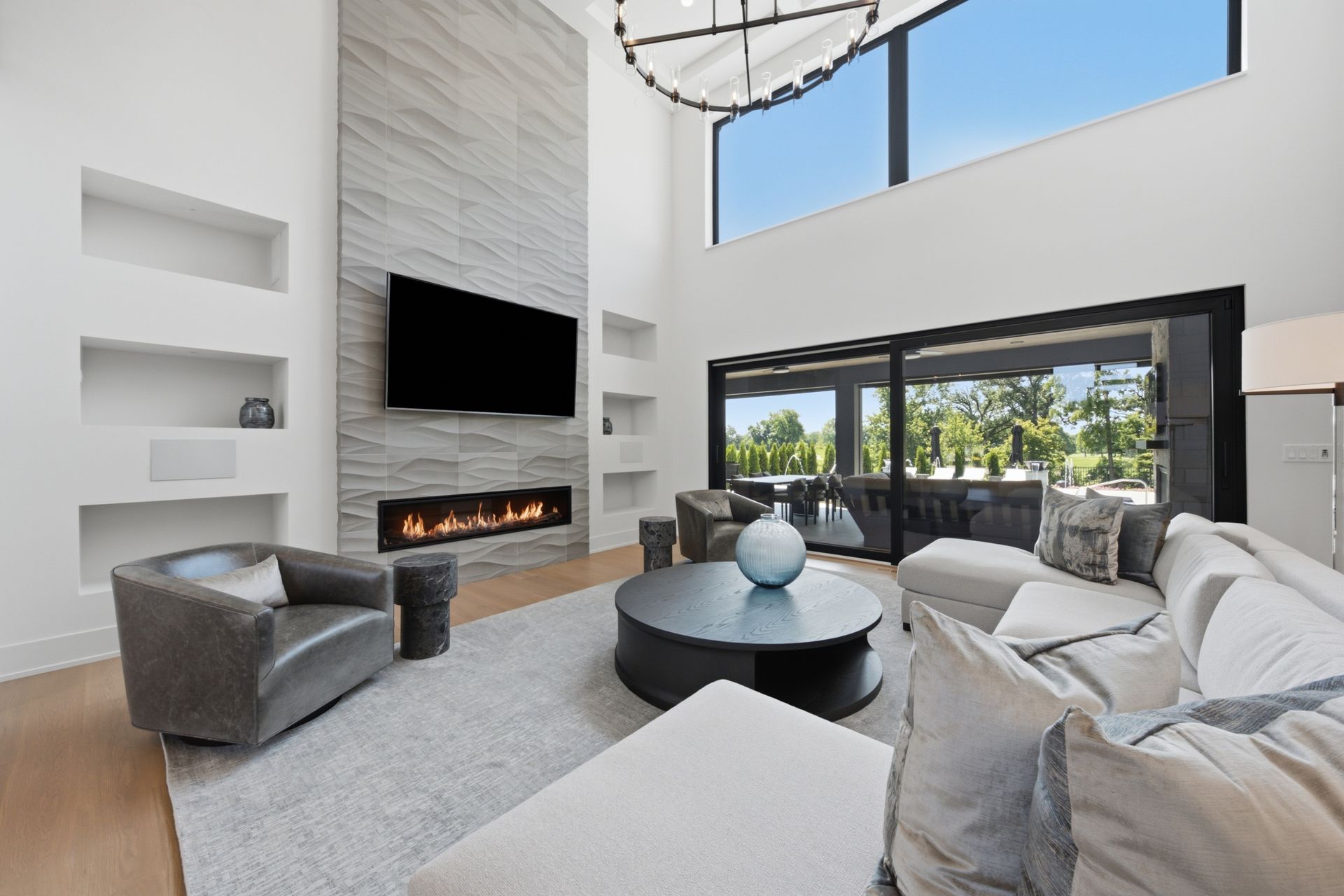 Modern living room with tall fireplace, wall-mounted TV, gray seating, and large windows overlooking greenery