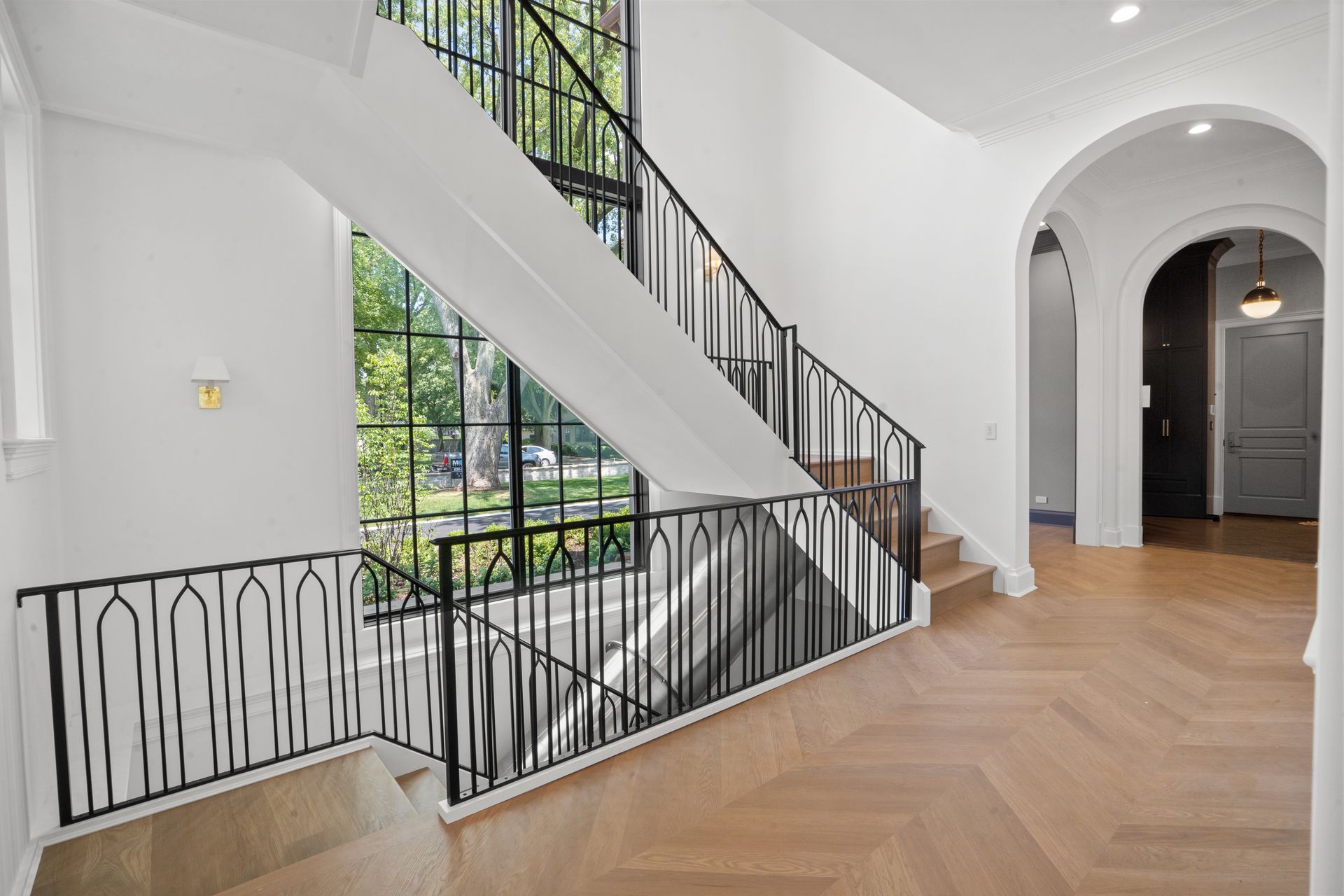Bright foyer with herringbone wood floors, staircase, black railing, and arched doorway in a modern home