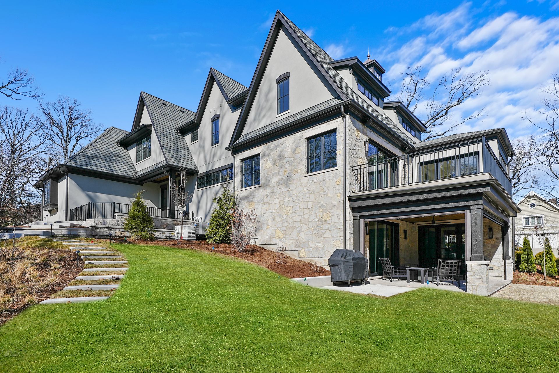 Large stone house with steep gables, wraparound deck, and green lawn under a blue sky.