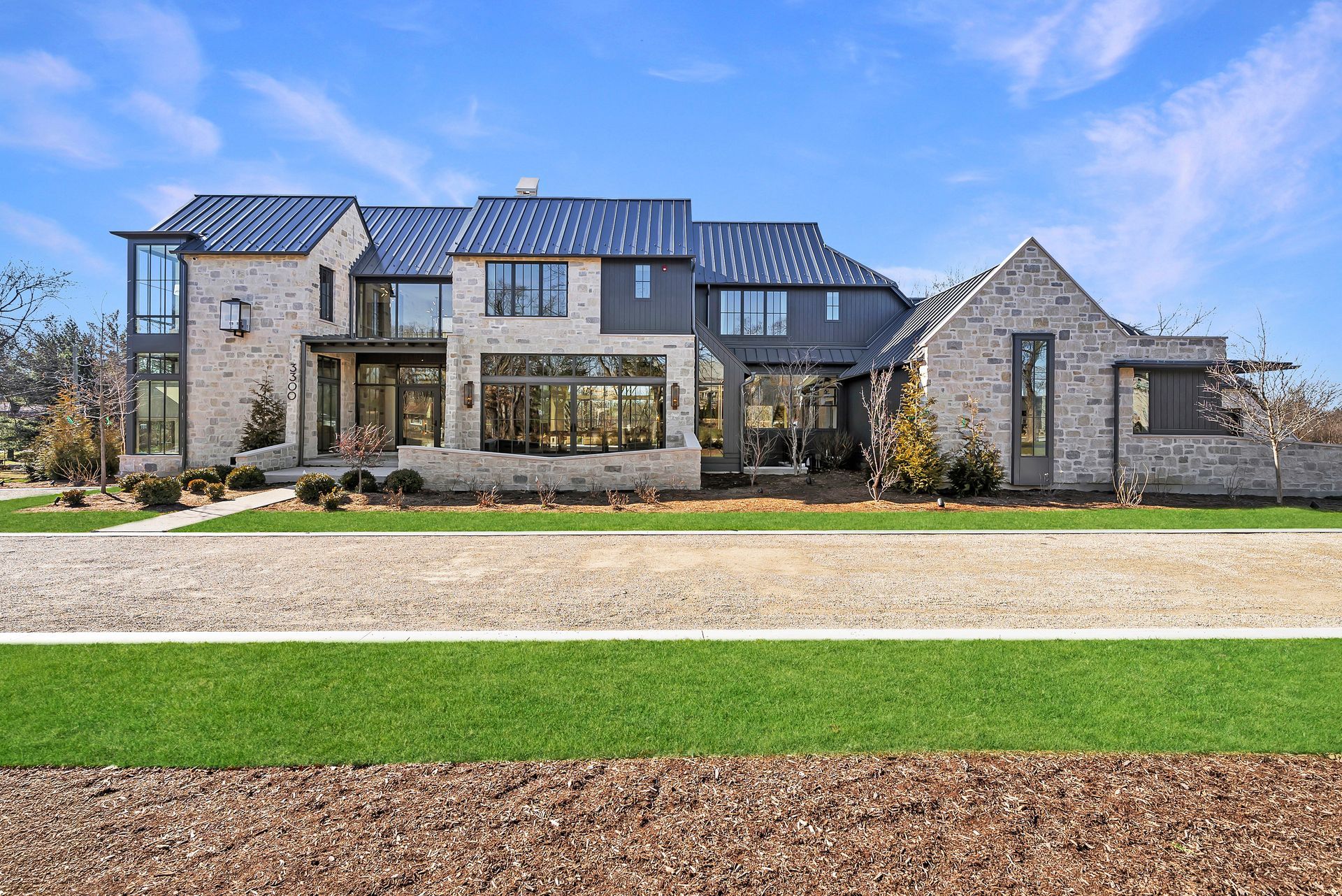Large modern stone house with dark metal roof, front lawn, and clear blue sky