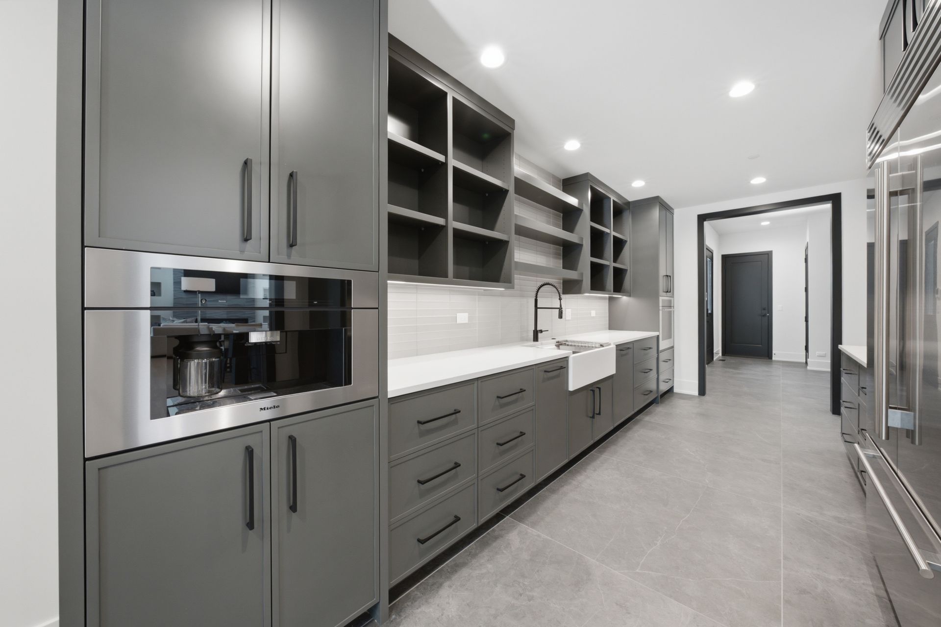 Modern gray kitchen with built-in appliances, long counter, and open shelving in a bright hallway.
