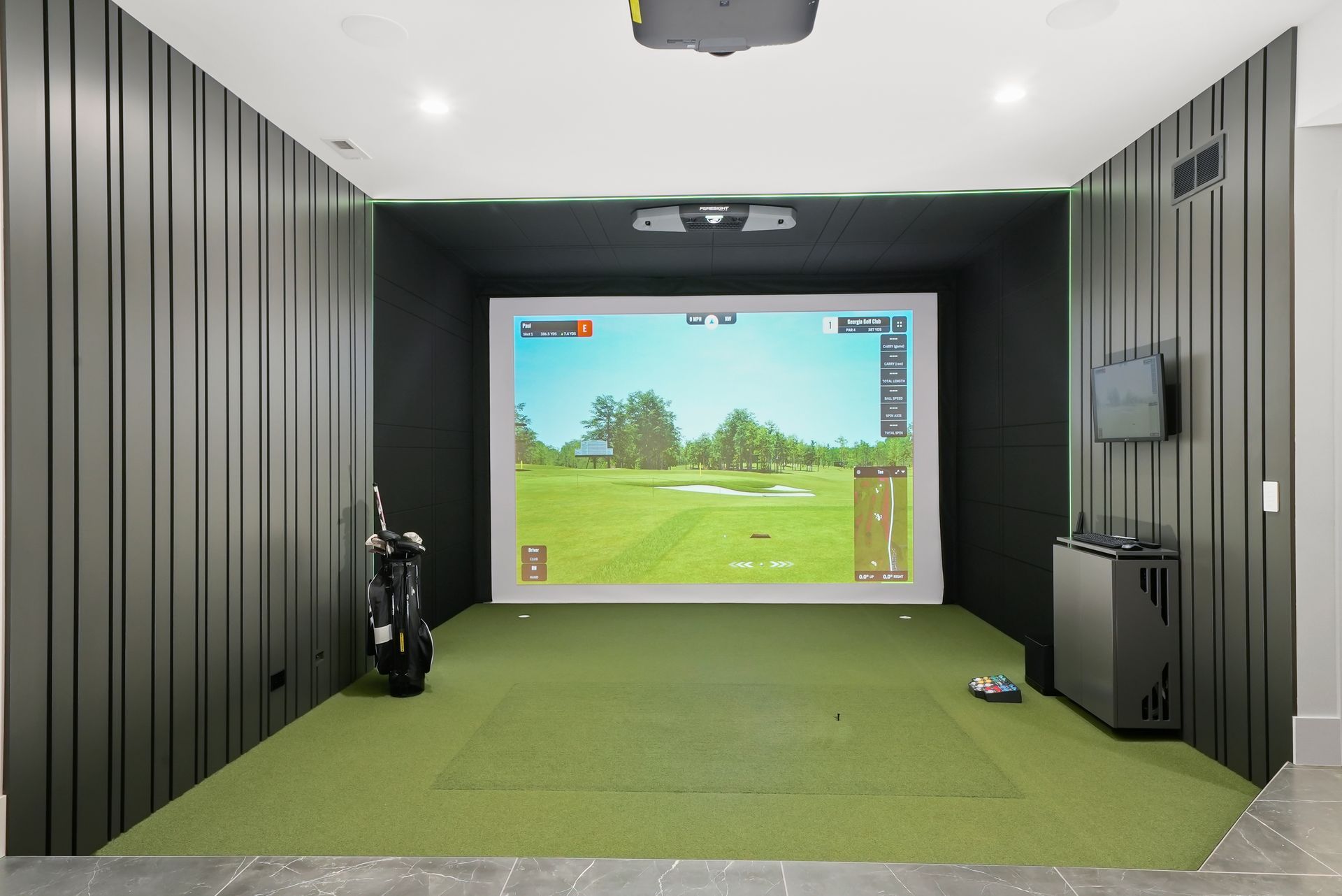 Indoor golf simulator room with green turf, projector screen, and black paneled walls