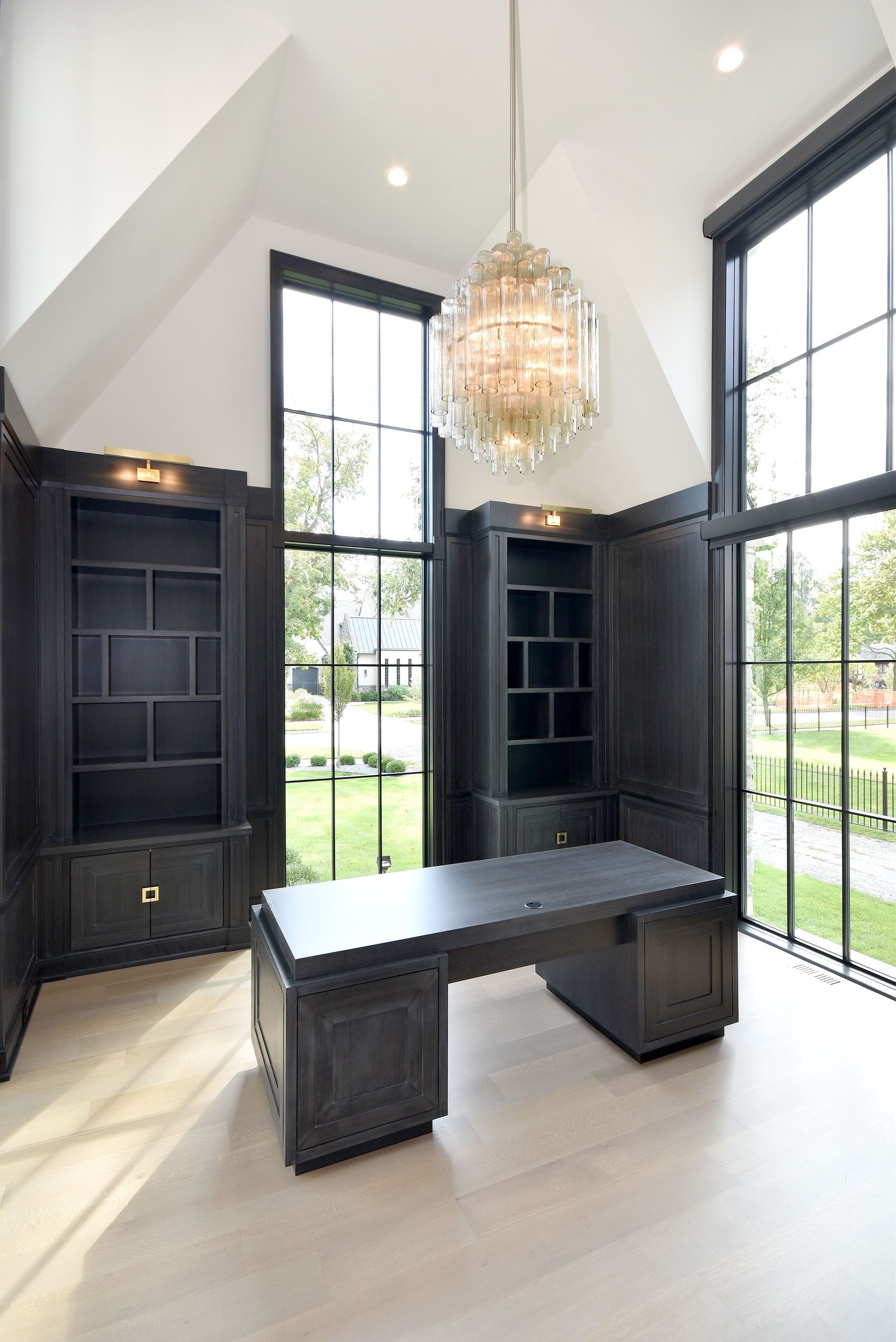 Modern office with dark built-in shelves, large windows, a black desk, and a chandelier.