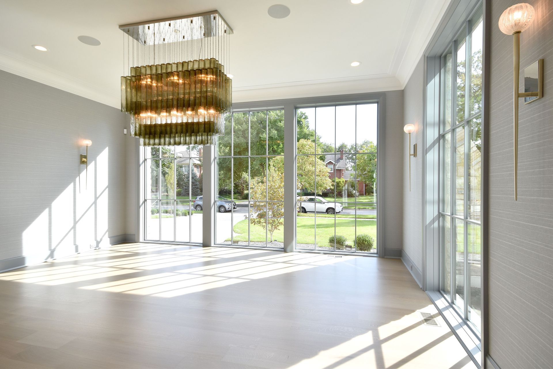 Bright modern empty room with large windows, glass doors, and a gold chandelier opening to a garden