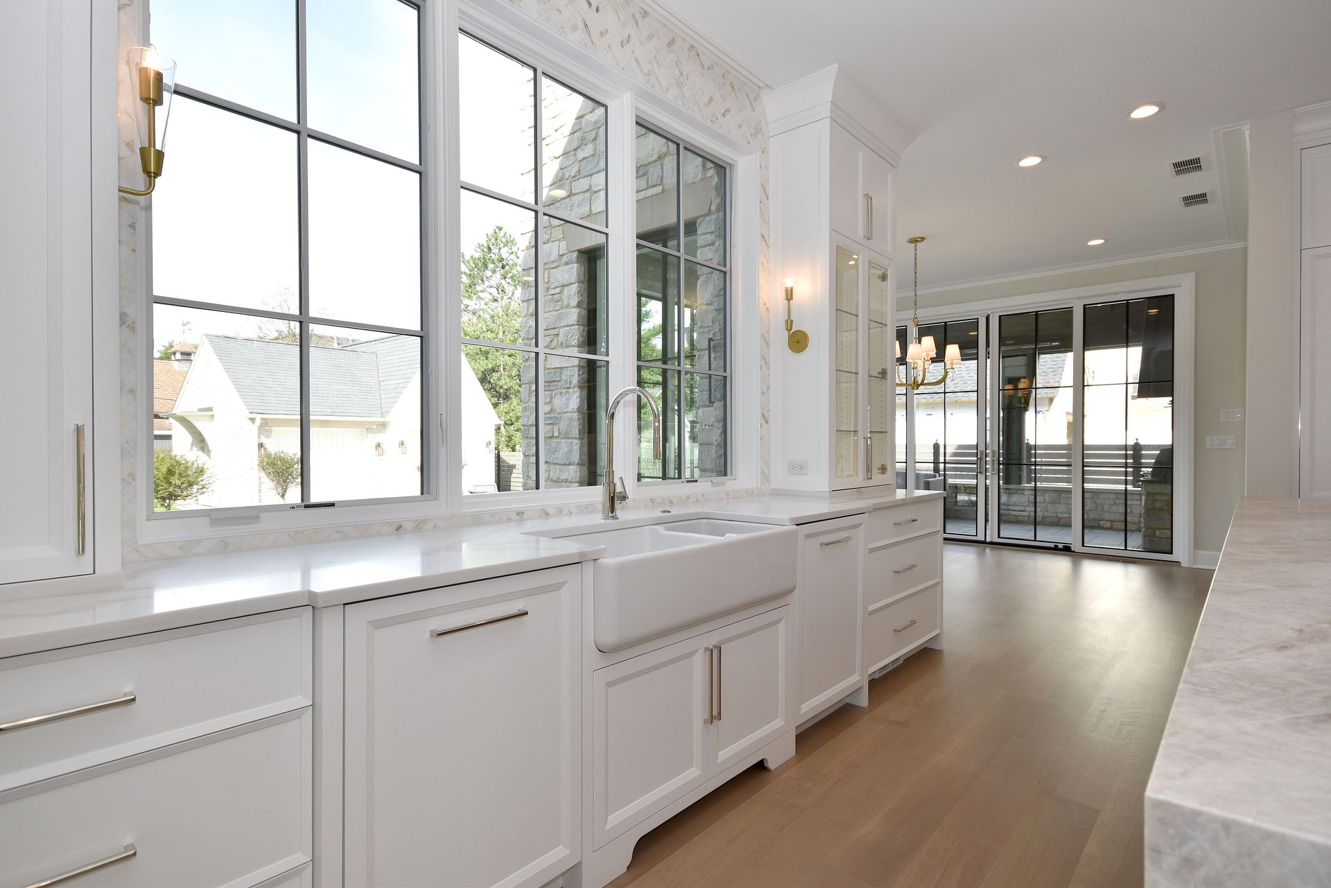Bright white kitchen with large windows, marble counters, and light wood floors leading to a glass door.