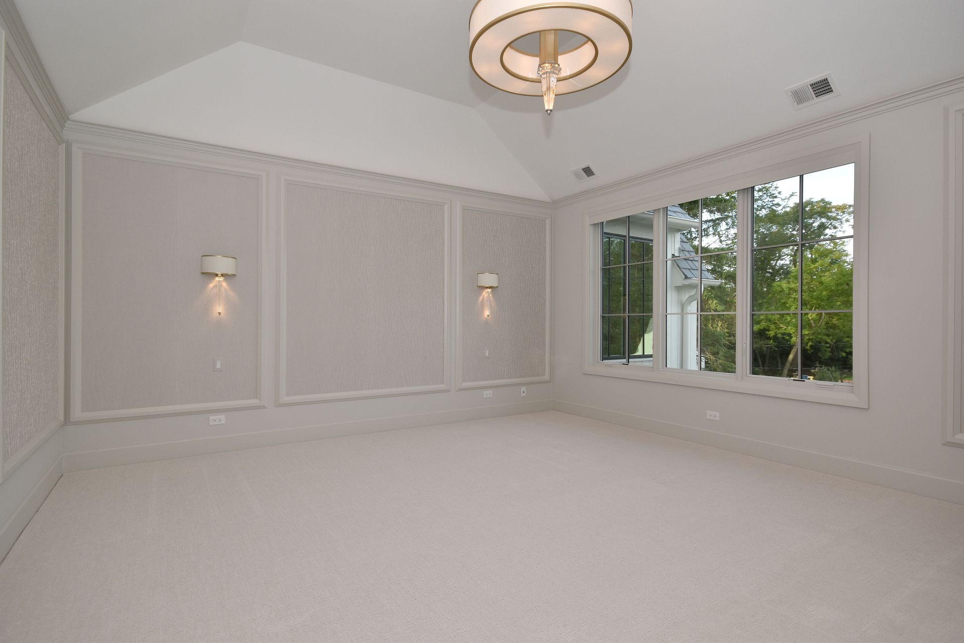Bright empty bedroom with white walls, chandelier, wall sconces, and large window overlooking greenery