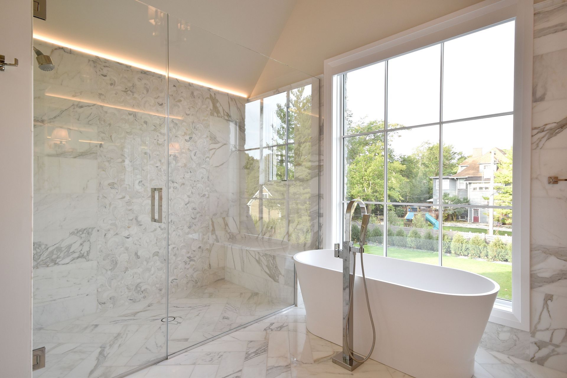 Bright marble bathroom with glass shower, freestanding tub, and large windows overlooking greenery.
