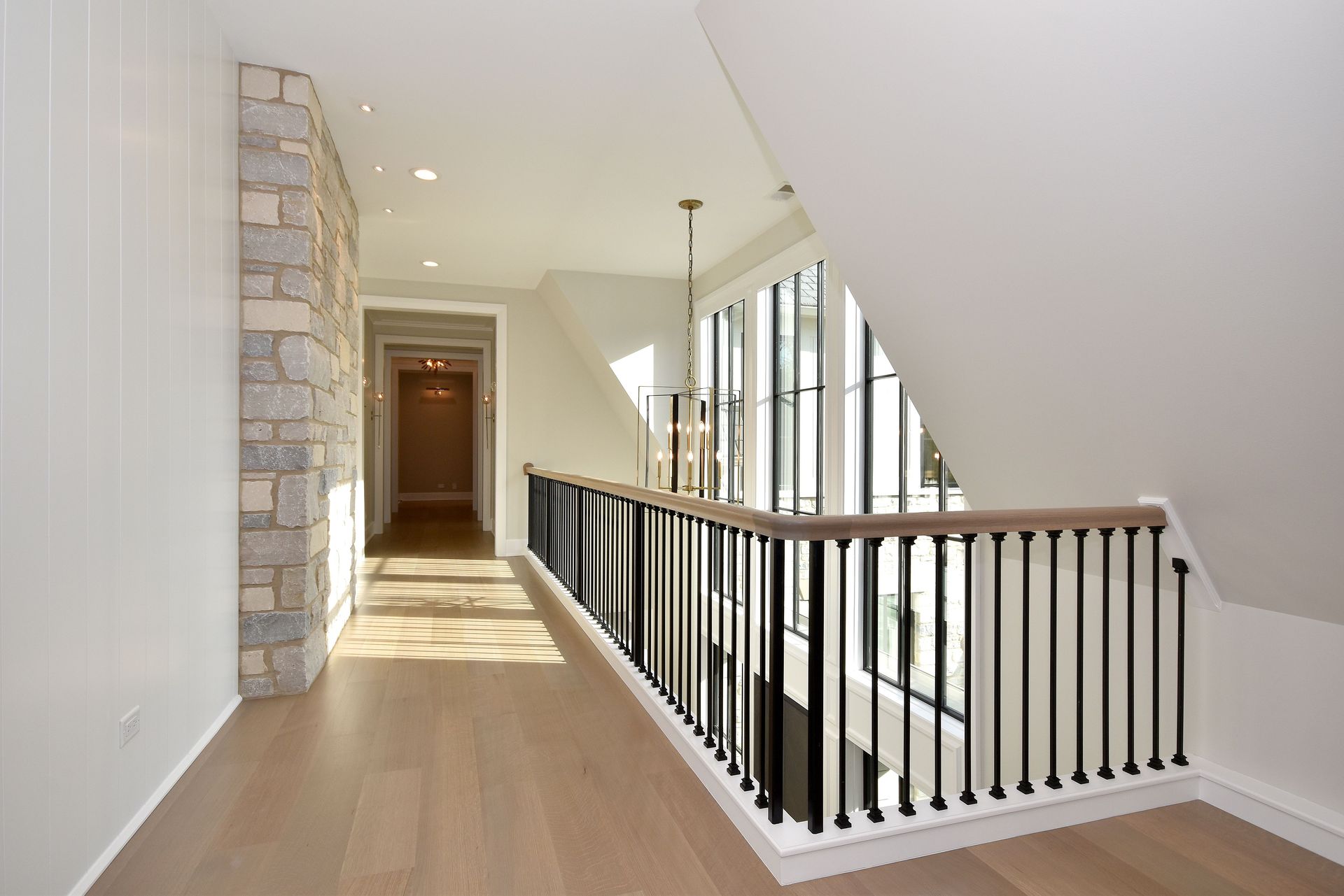 Bright upstairs hallway with stone accent wall, black railing, and sloped ceiling by large windows.