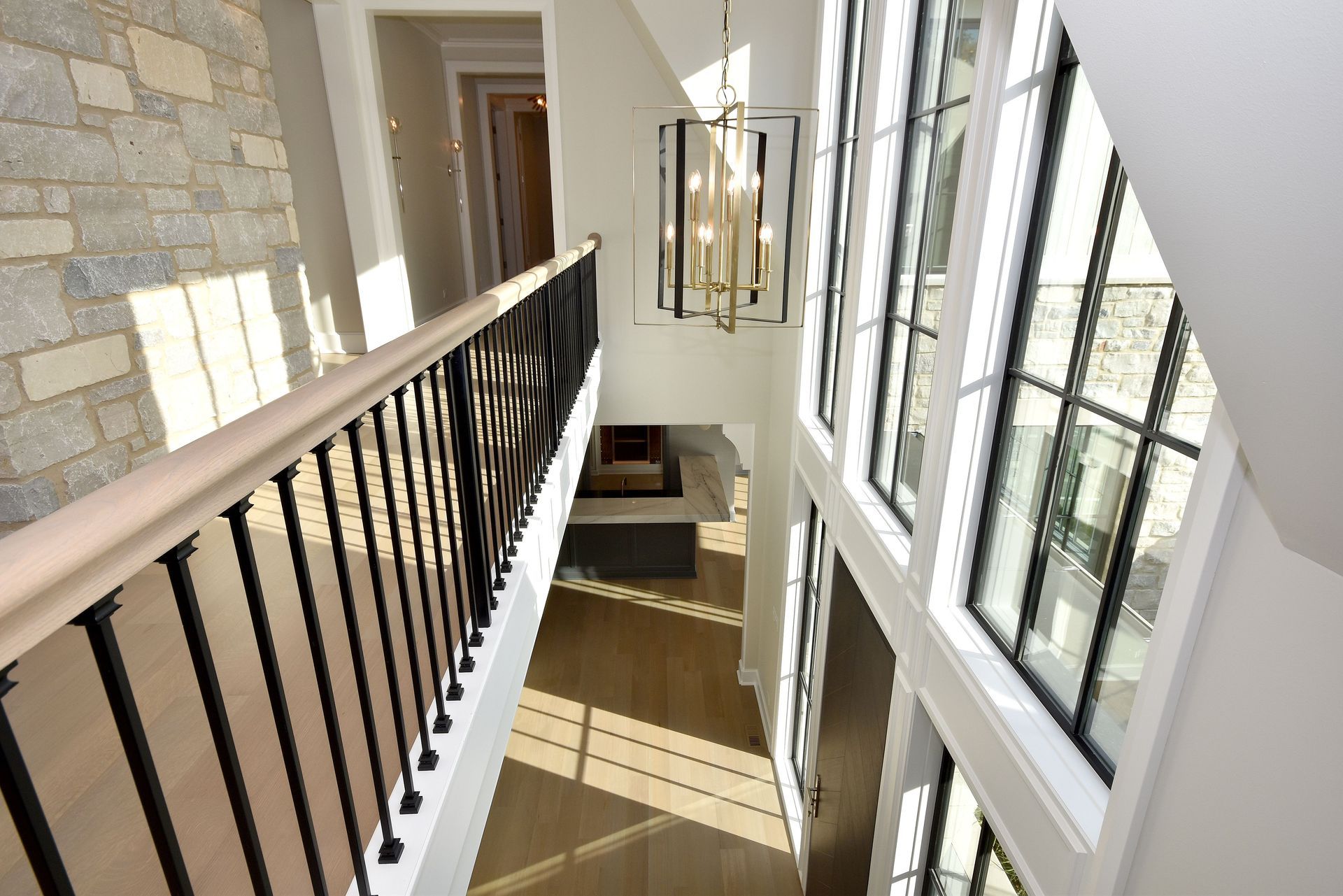 Bright interior balcony hallway with black railing, stone wall, and tall windows overlooking a lower floor.