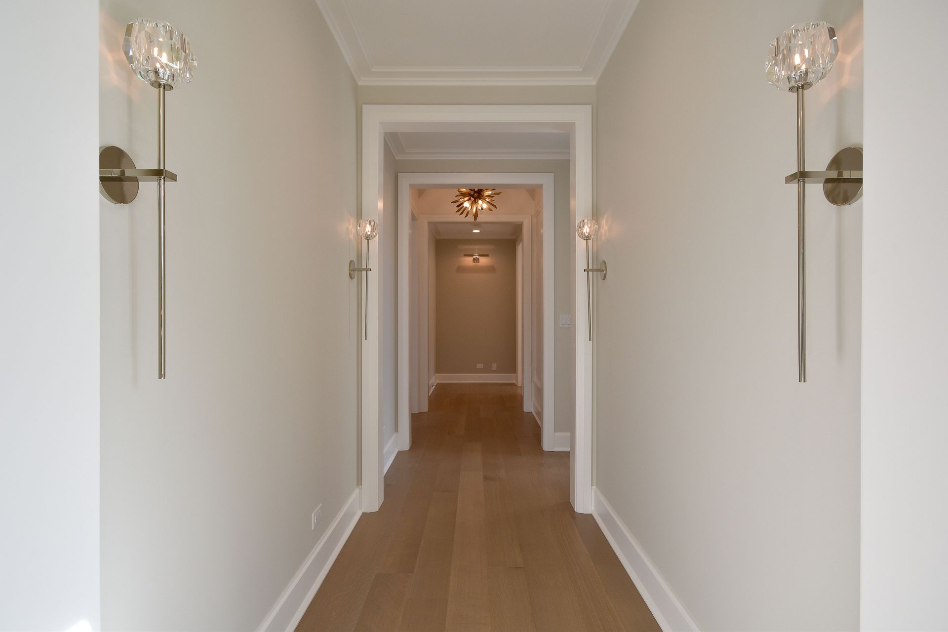 Bright white hallway with wood floors, wall sconces, and a doorway at the end.