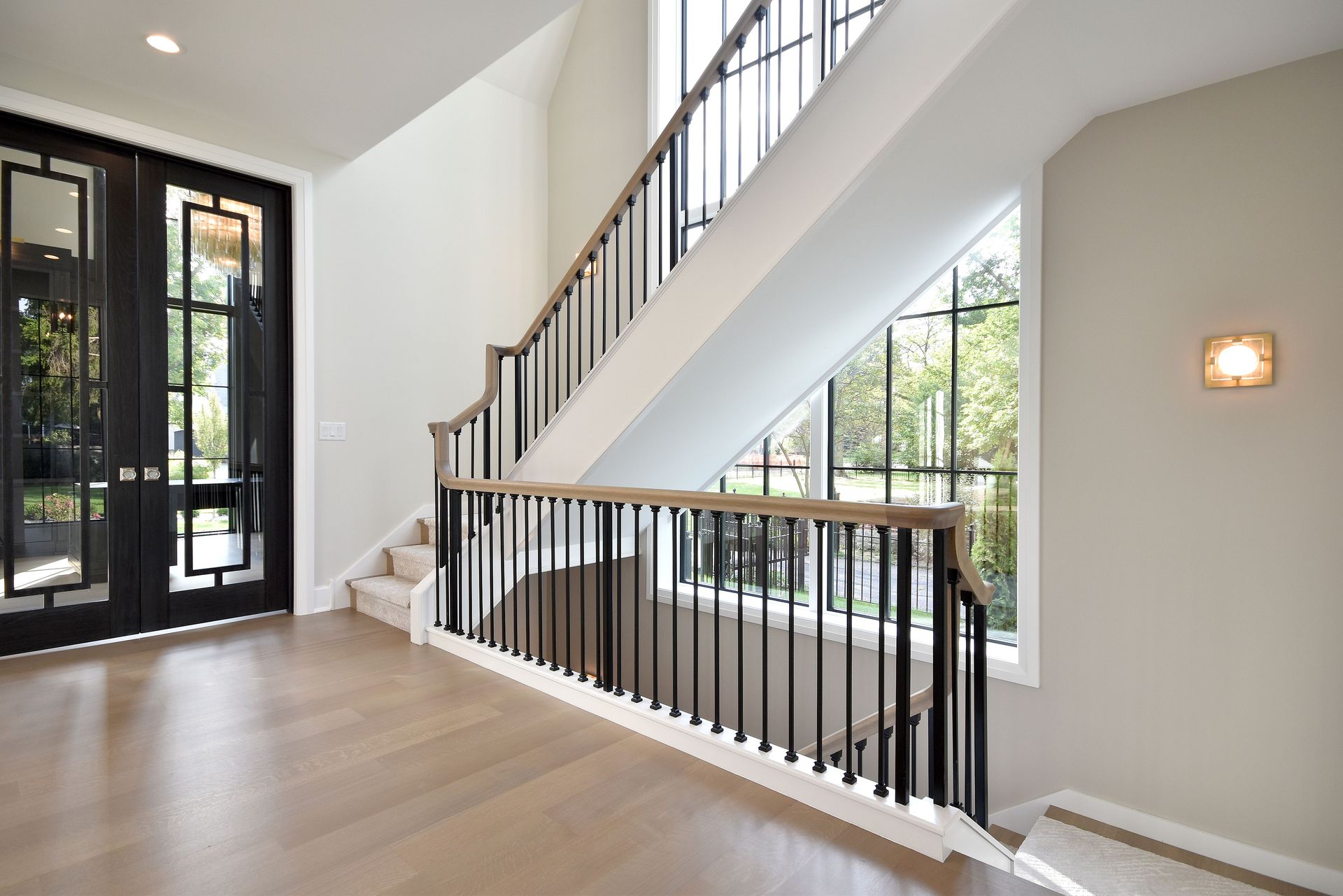 Bright foyer with staircase, black railing, light wood floors, and large windows.