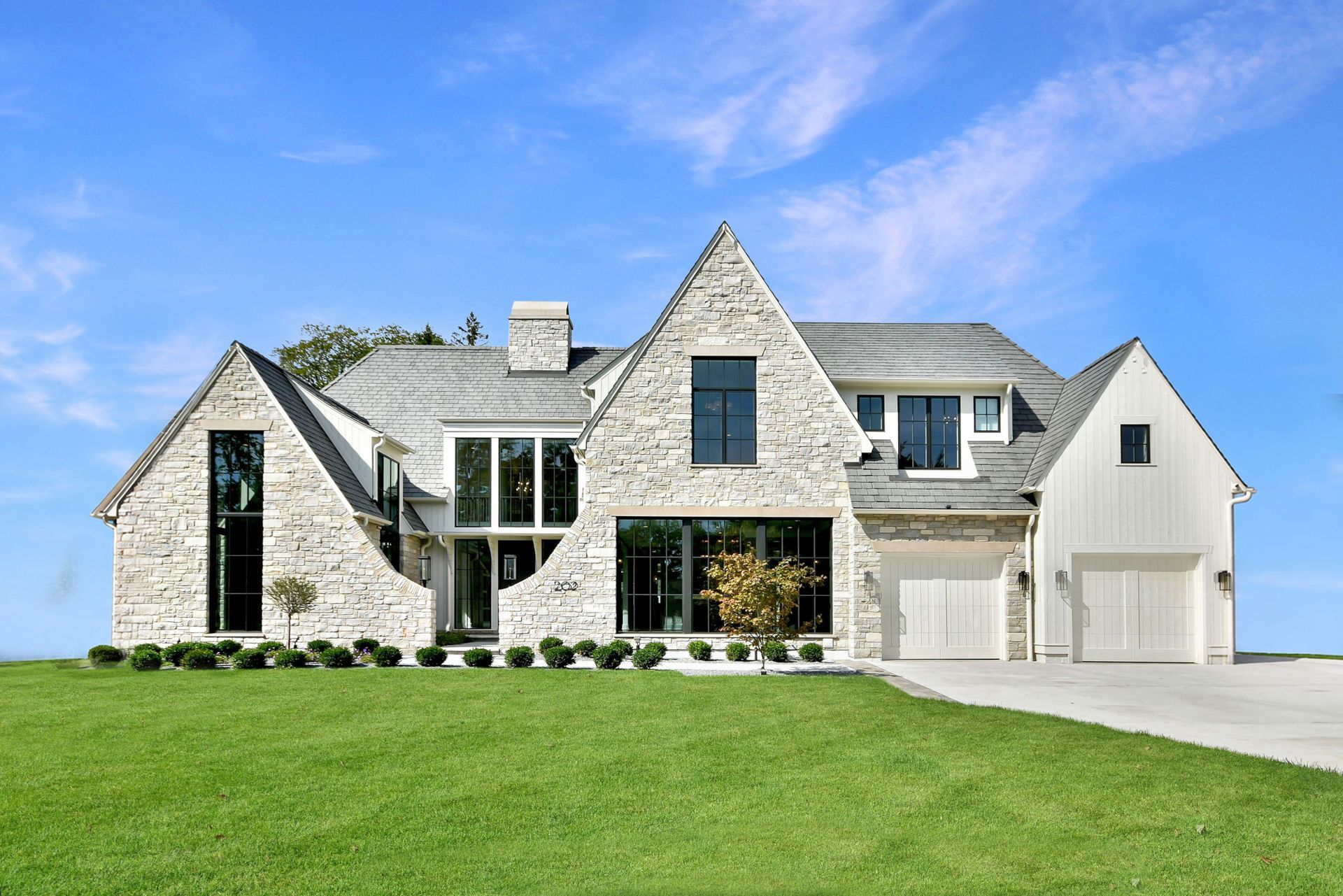 Large stone suburban house with a gray roof, white garage, and a green lawn under a blue sky
