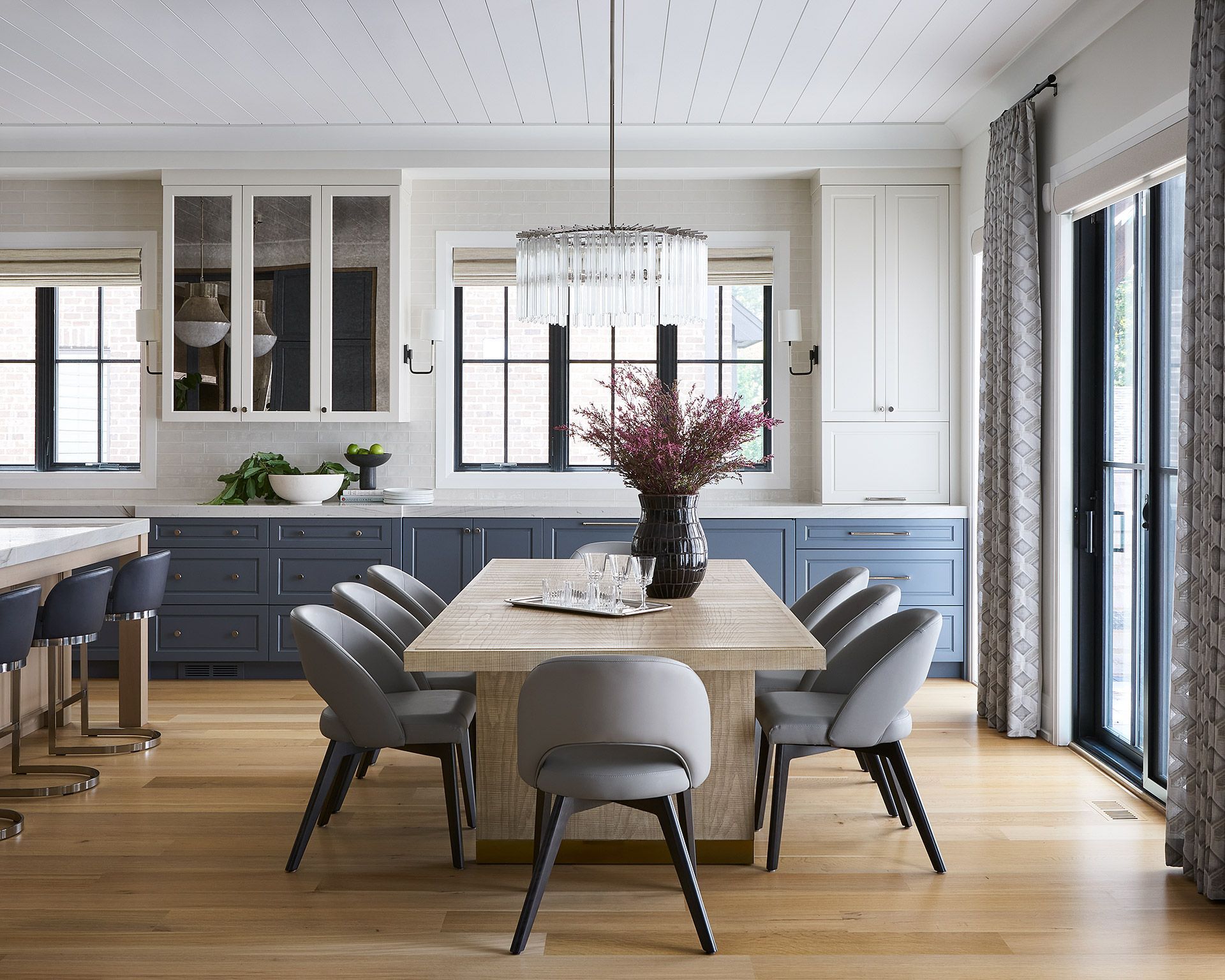 Modern dining room with a wooden table, gray chairs, and blue cabinets beside large windows.