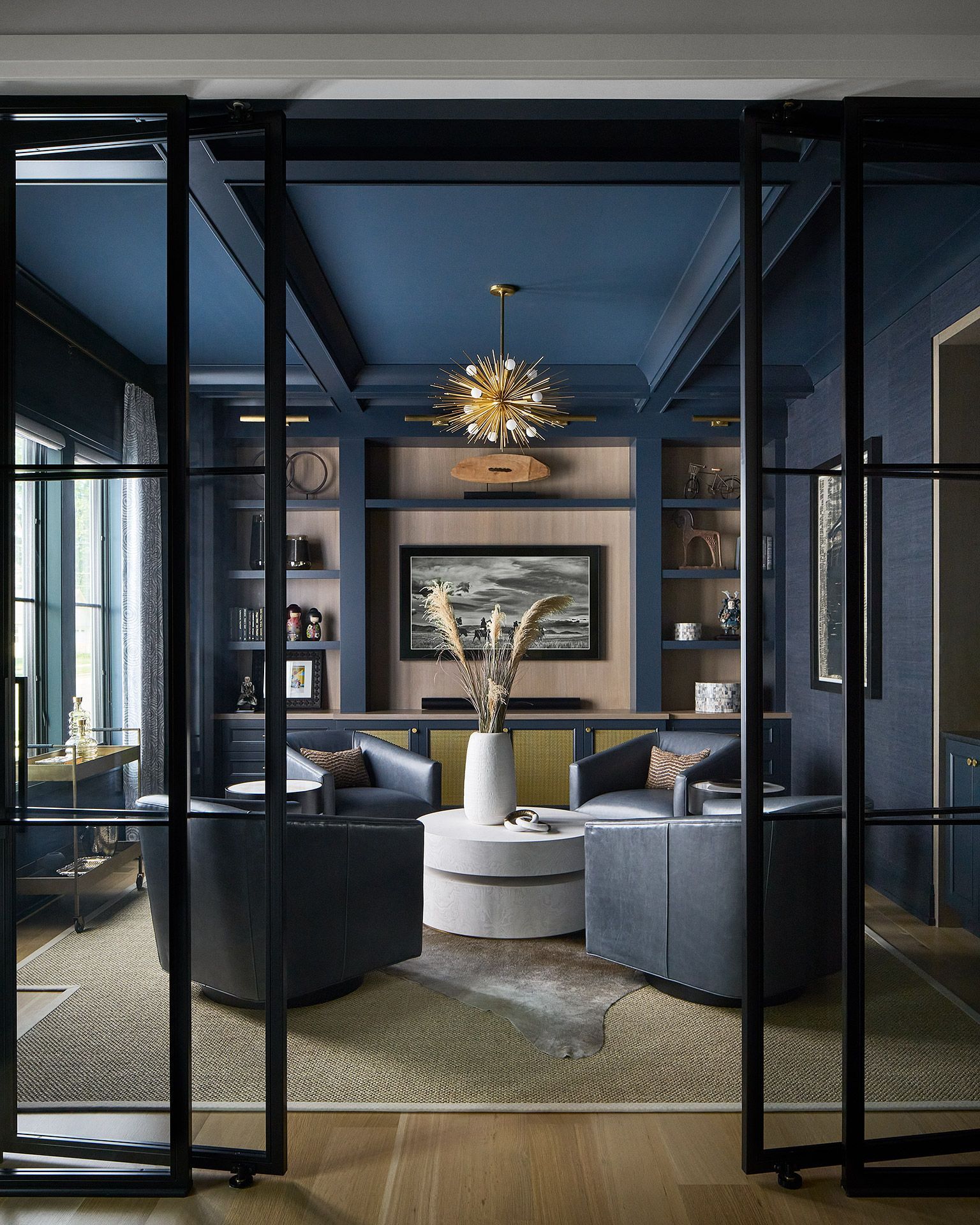 Modern navy lounge with glass doors, built-in shelves, round coffee table, and gold chandelier
