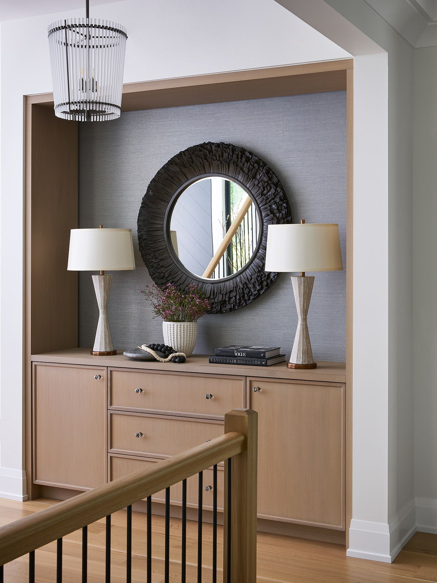 Built-in hallway cabinet with two lamps, round mirror, and wood rail in a neutral interior.