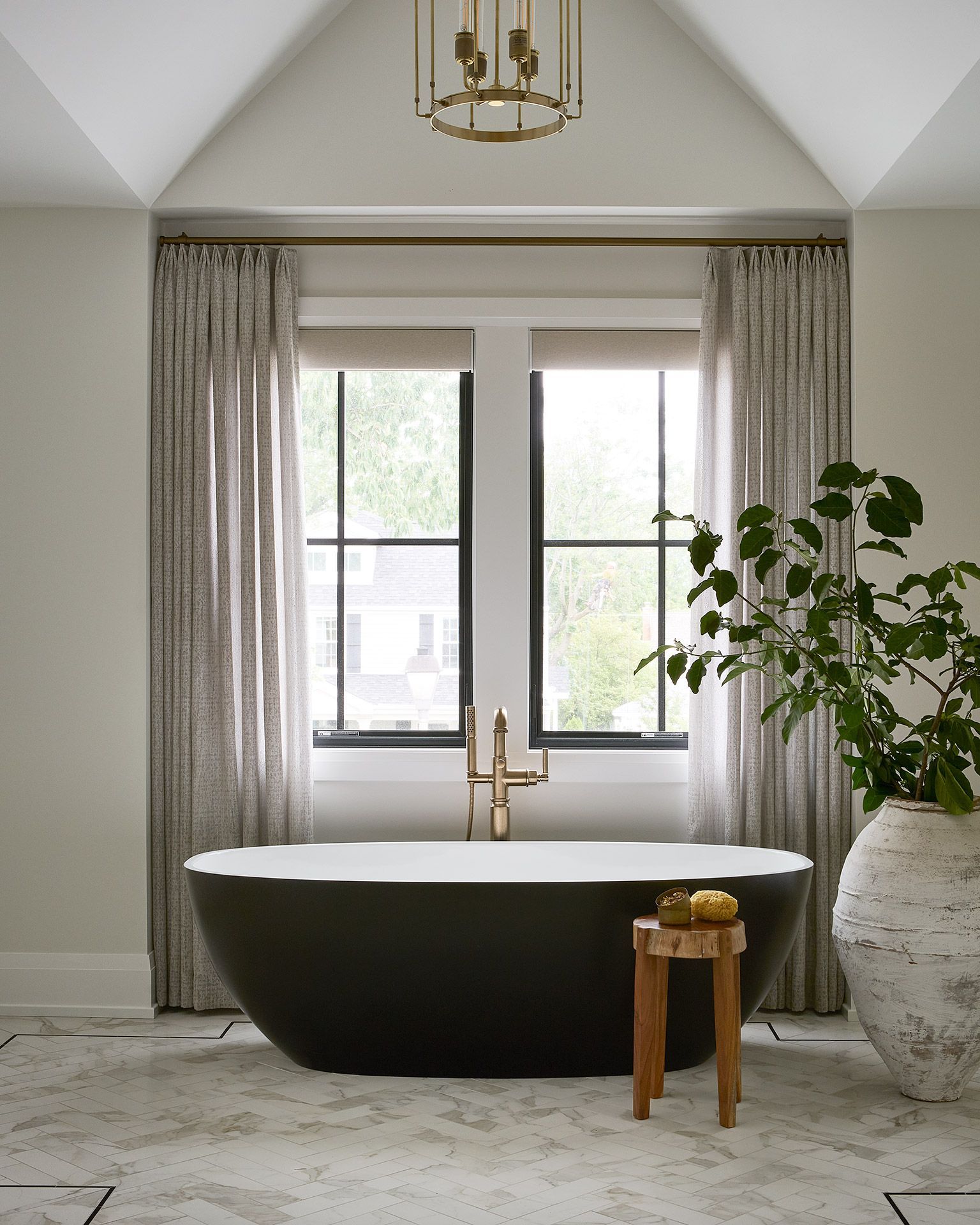 Modern bathroom with a freestanding black tub, window, curtains, stool, and large potted plant.