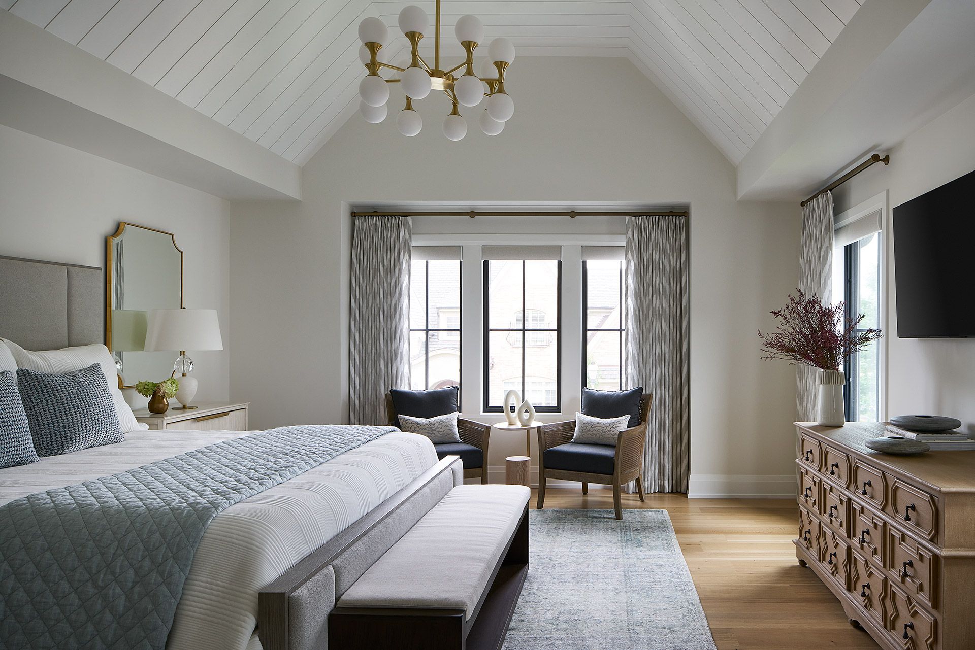 Bright bedroom with vaulted ceiling, gray bed, window seats, and a carved dresser by a window.